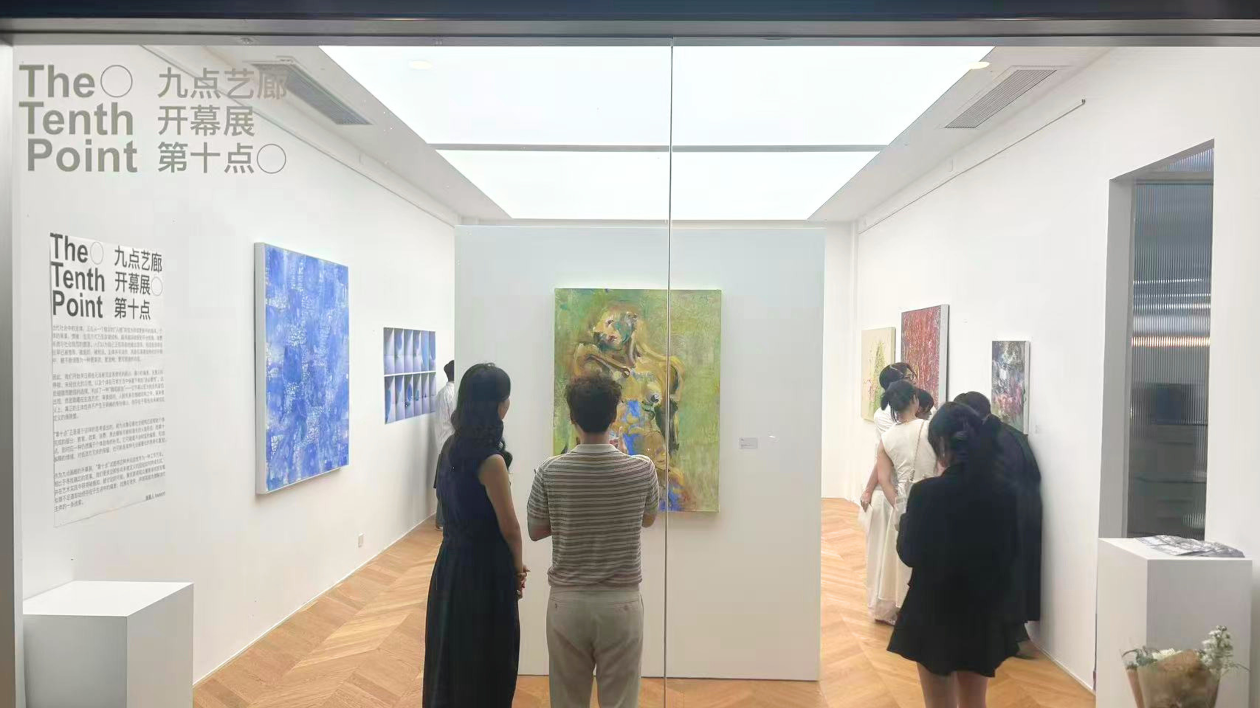 People viewing paintings in an art gallery with white walls and wooden floors, glass doors, and ceiling lights.