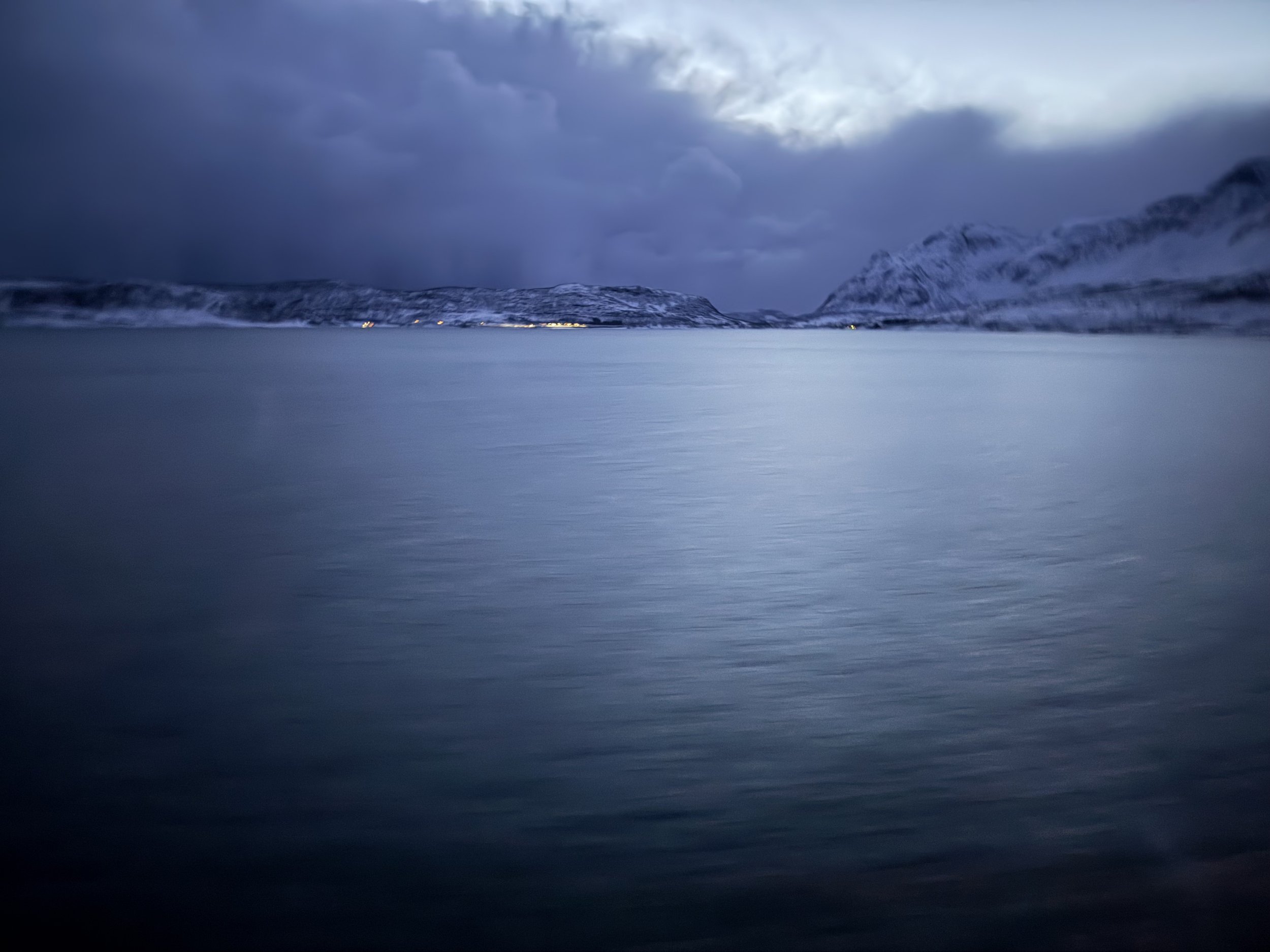 A snowy landscape with mountains under a dark, cloudy sky, and a body of water in the foreground.