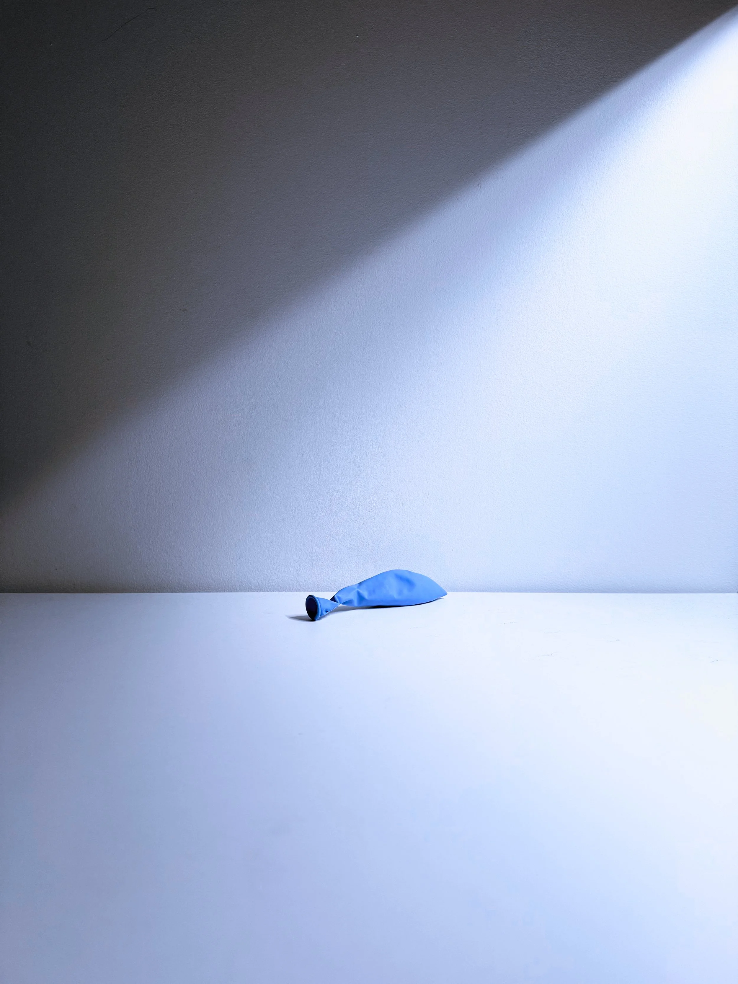 A deflated blue balloon lying on a white surface with a stark shadow cast on a light gray wall behind it.