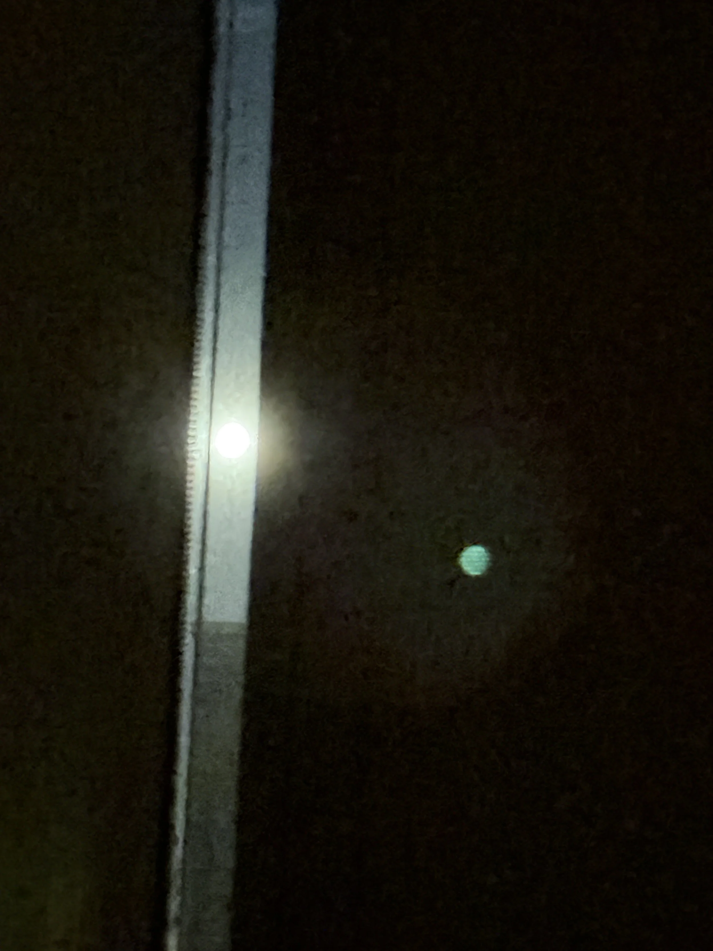 Nighttime scene showing a tall streetlight illuminating a dark street, with some distant smaller lights visible in the background.