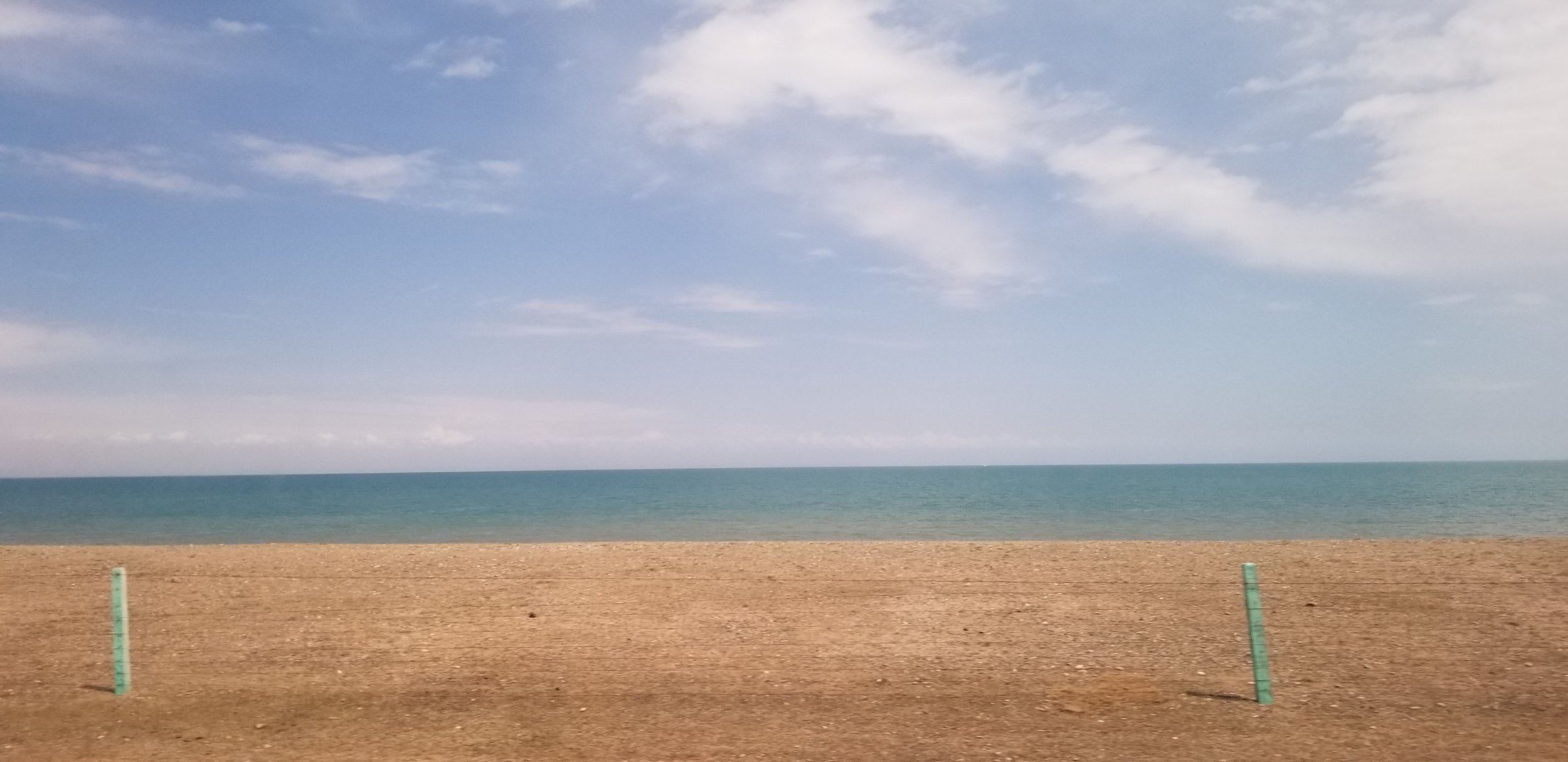 A beach scene with a sandy shoreline, calm turquoise ocean water, and a partly cloudy blue sky.