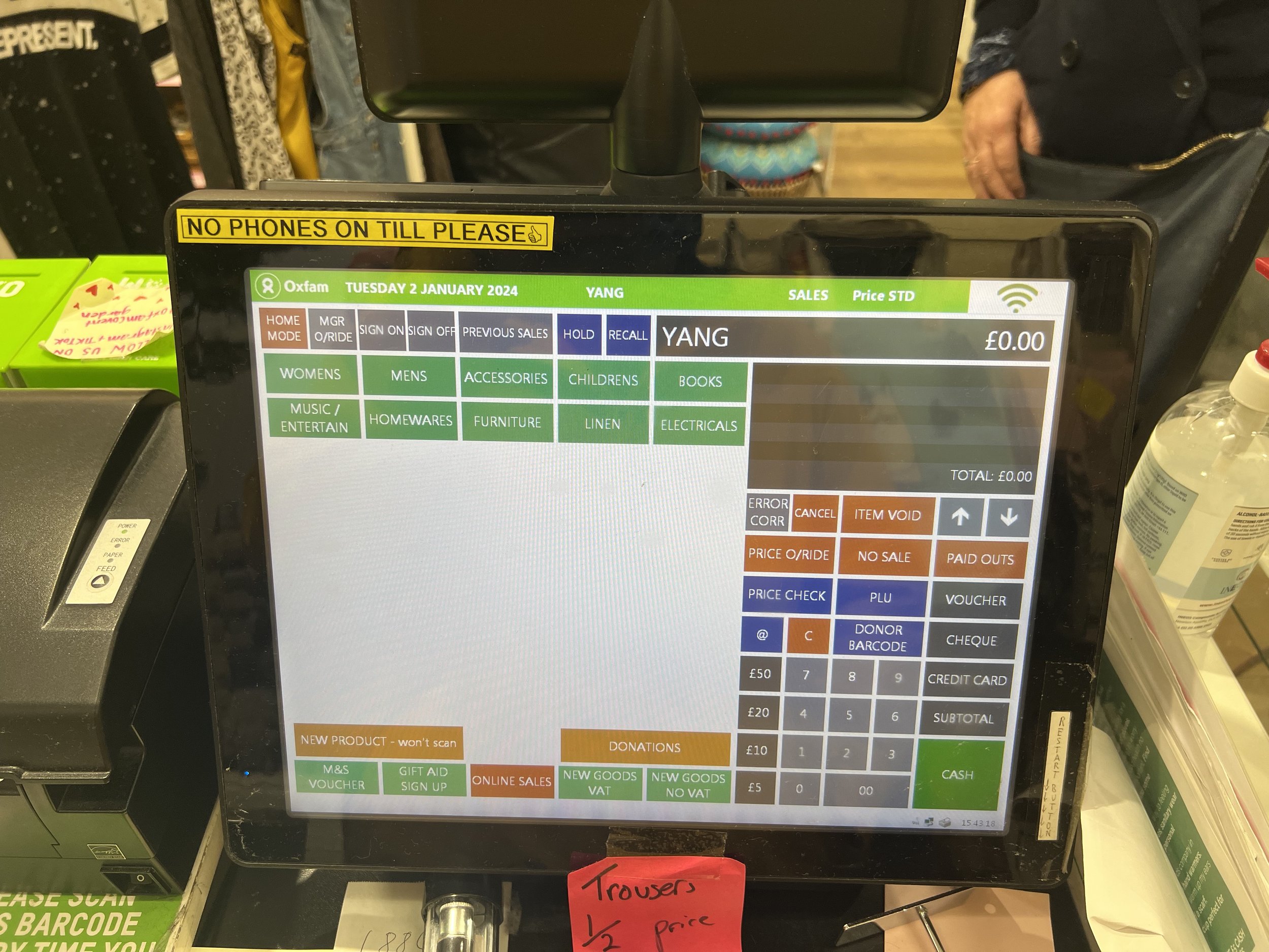 Point-of-sale touchscreen display showing date, transaction details, and various menu options including categories like women's, men's, accessories, children's, books, music, entertainment, homewares, furniture, linen, electricals, and donation optio