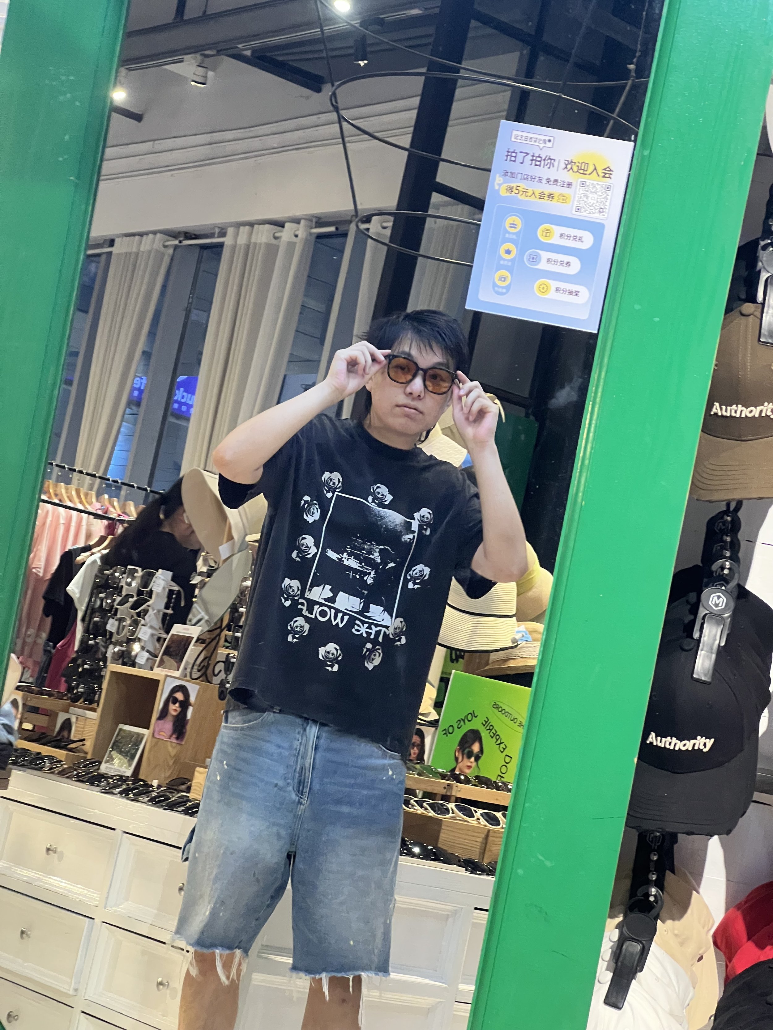 A person taking a mirror selfie in a clothing store wearing a black T-shirt with white roses design and denim shorts. The store has curtains, sunglasses, and displayed accessories.