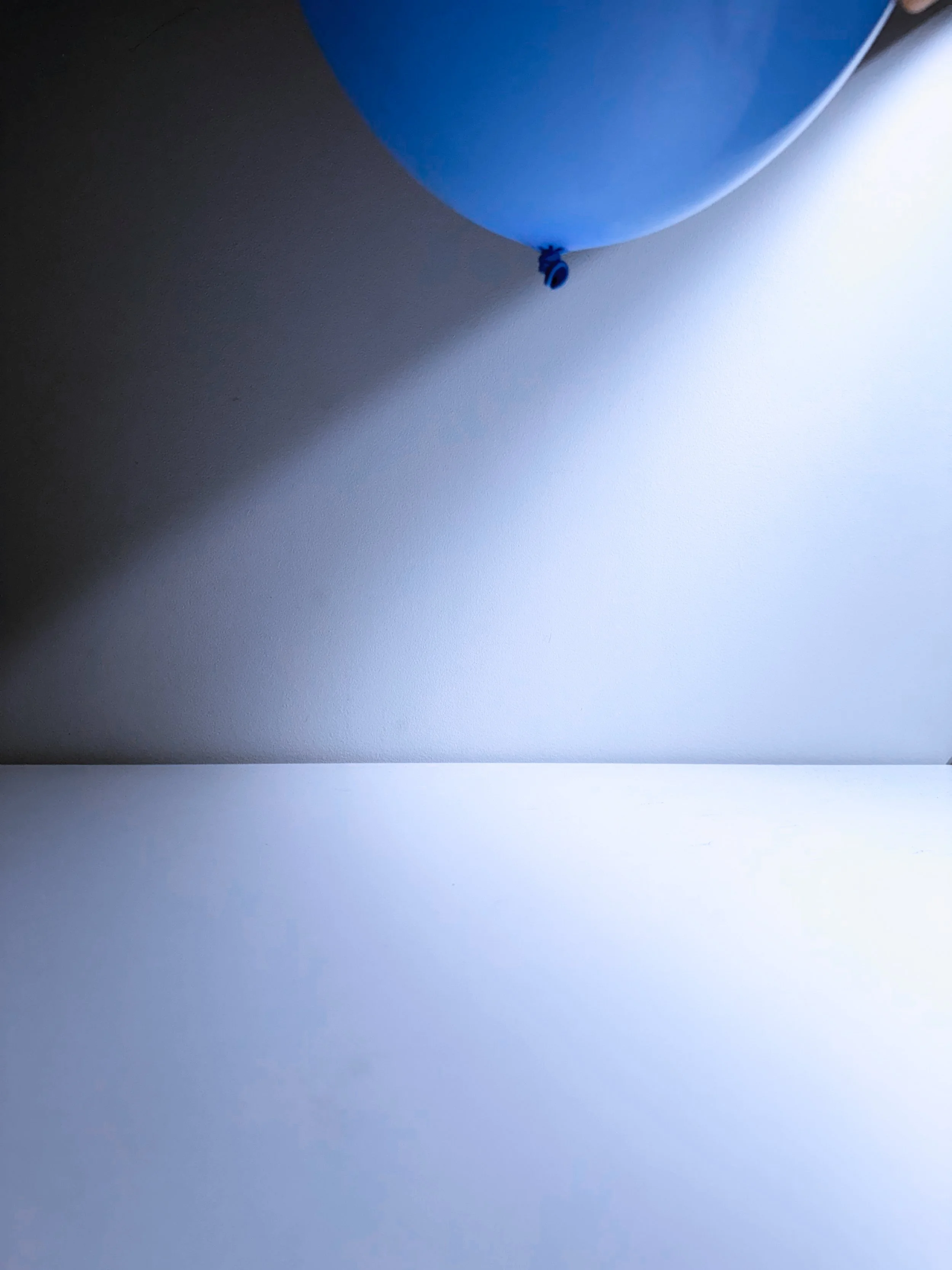 Blue balloon partially visible, tied with a small blue knot, hanging against a white wall, with a shadow cast on the surface.