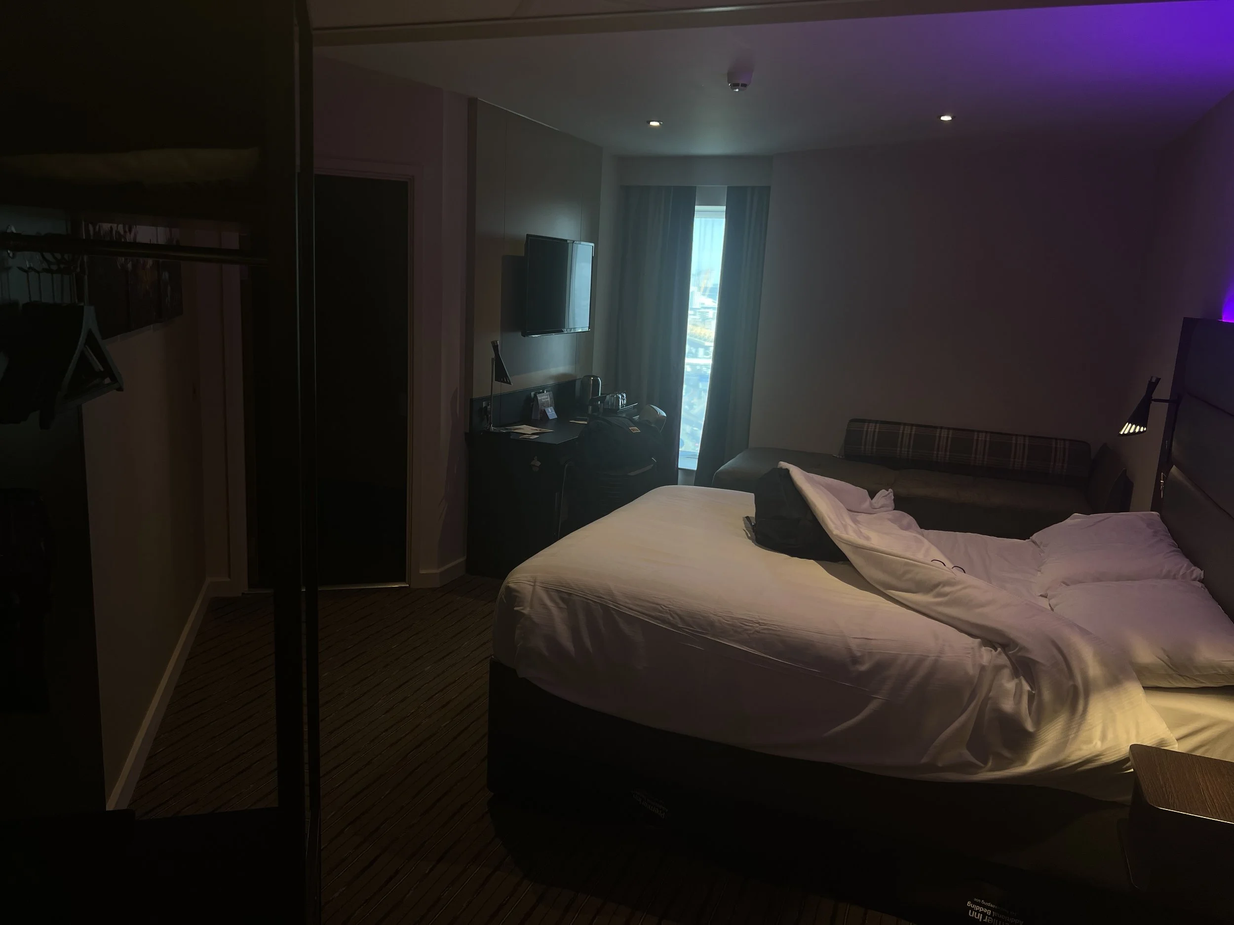 A hotel room with a neatly made bed, a window with curtains, a wall-mounted television, and some luggage and items on a black desk.