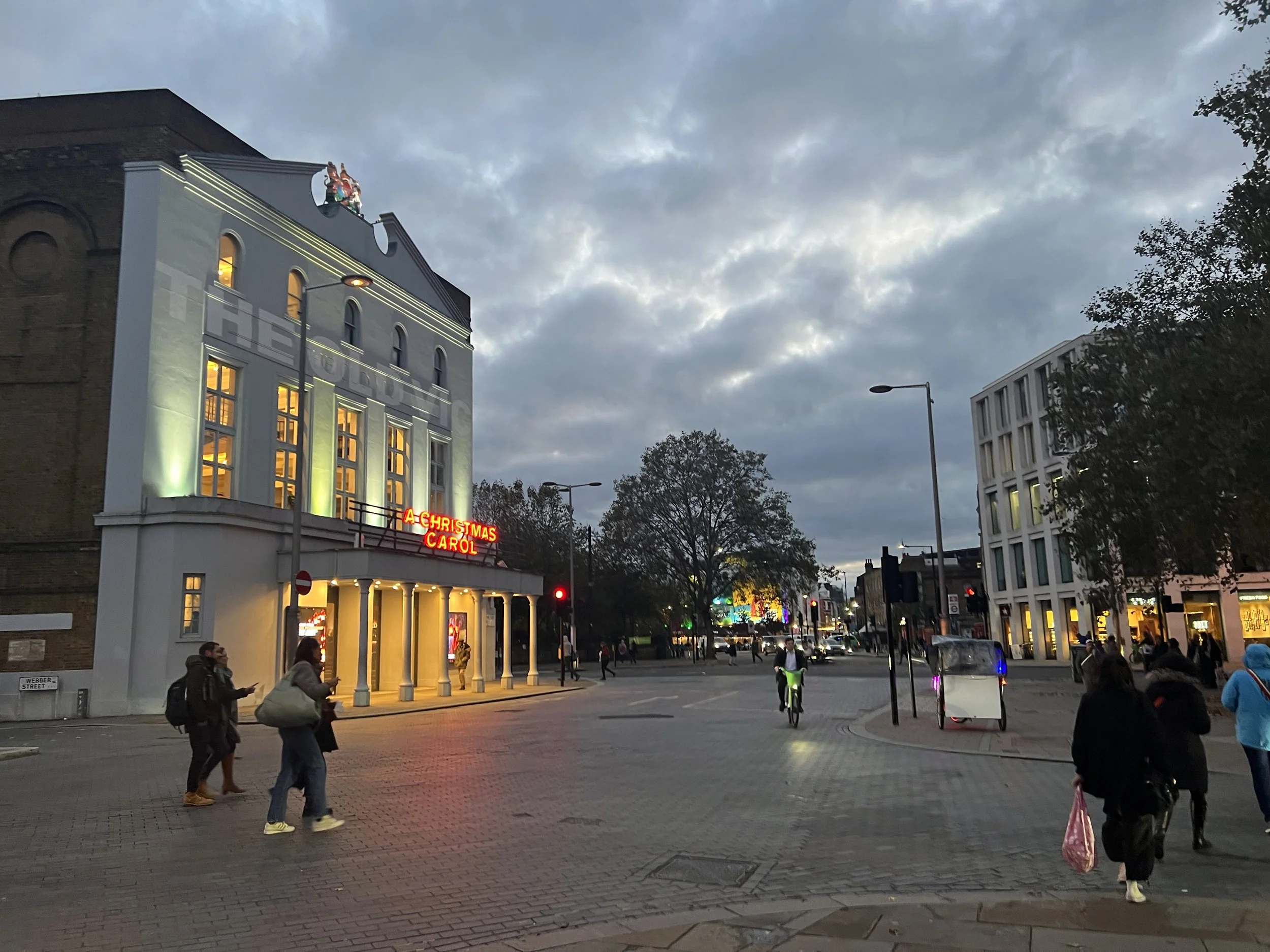 People walking and riding bikes on a city street in the evening, with illuminated buildings and a cloudy sky overhead.