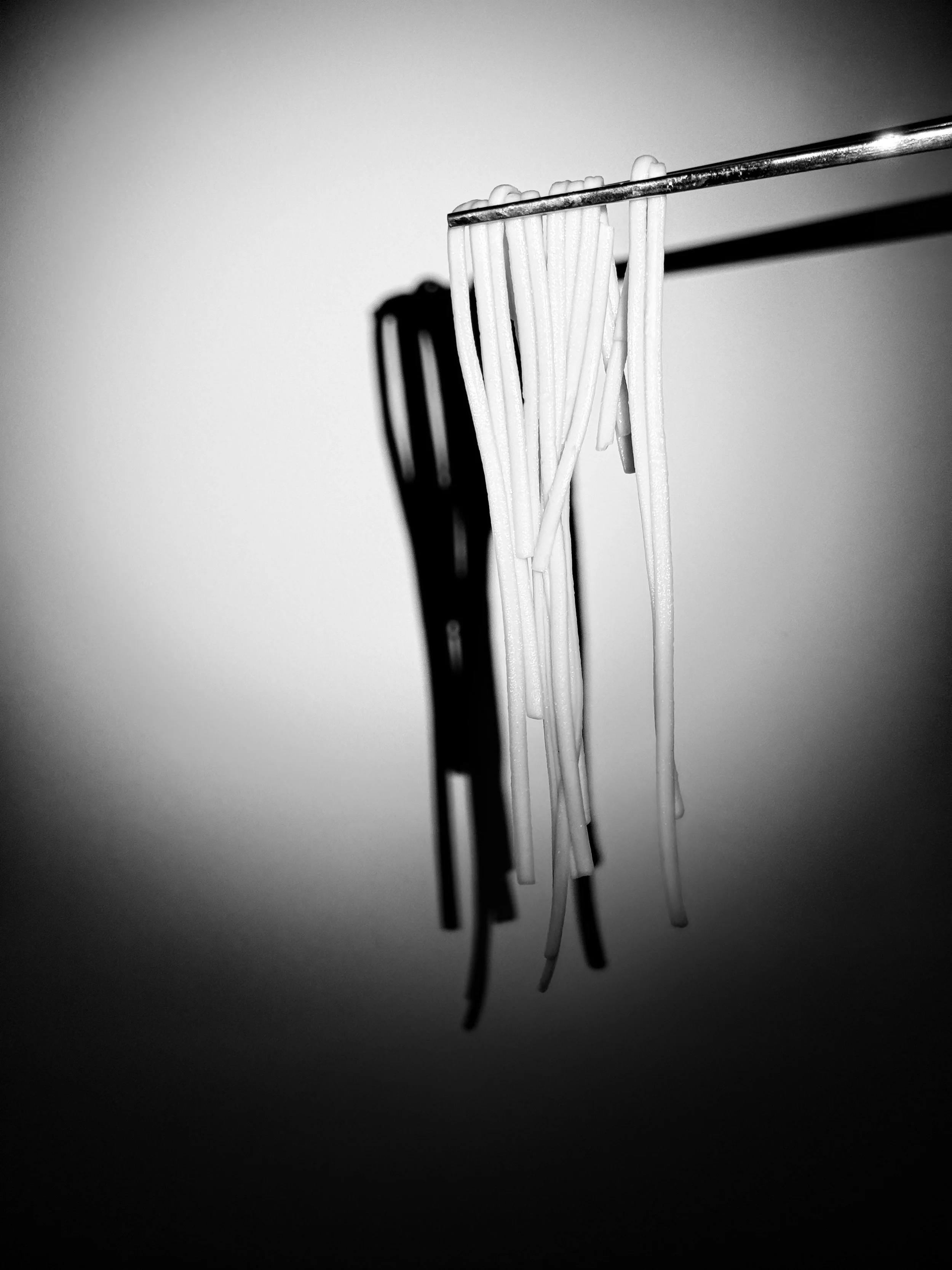 Black and white photograph of a few strands of spaghetti noodles hanging from a metal tool, with stark shadows cast on a plain background.