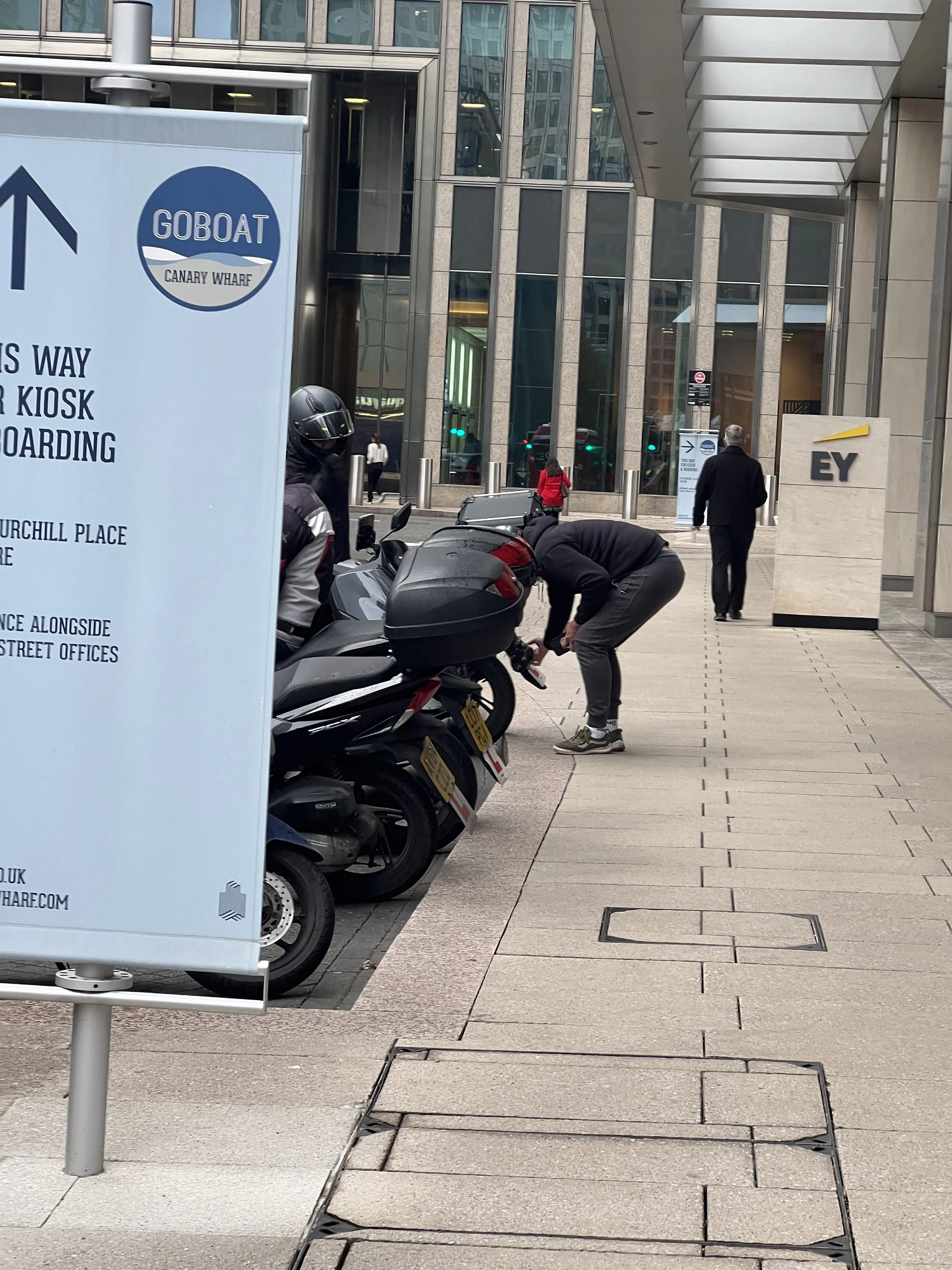 Sidewalk scene in an urban area with several parked scooters, a person with a helmet, two people walking, and signs for Canary Wharf and EY in the background.
