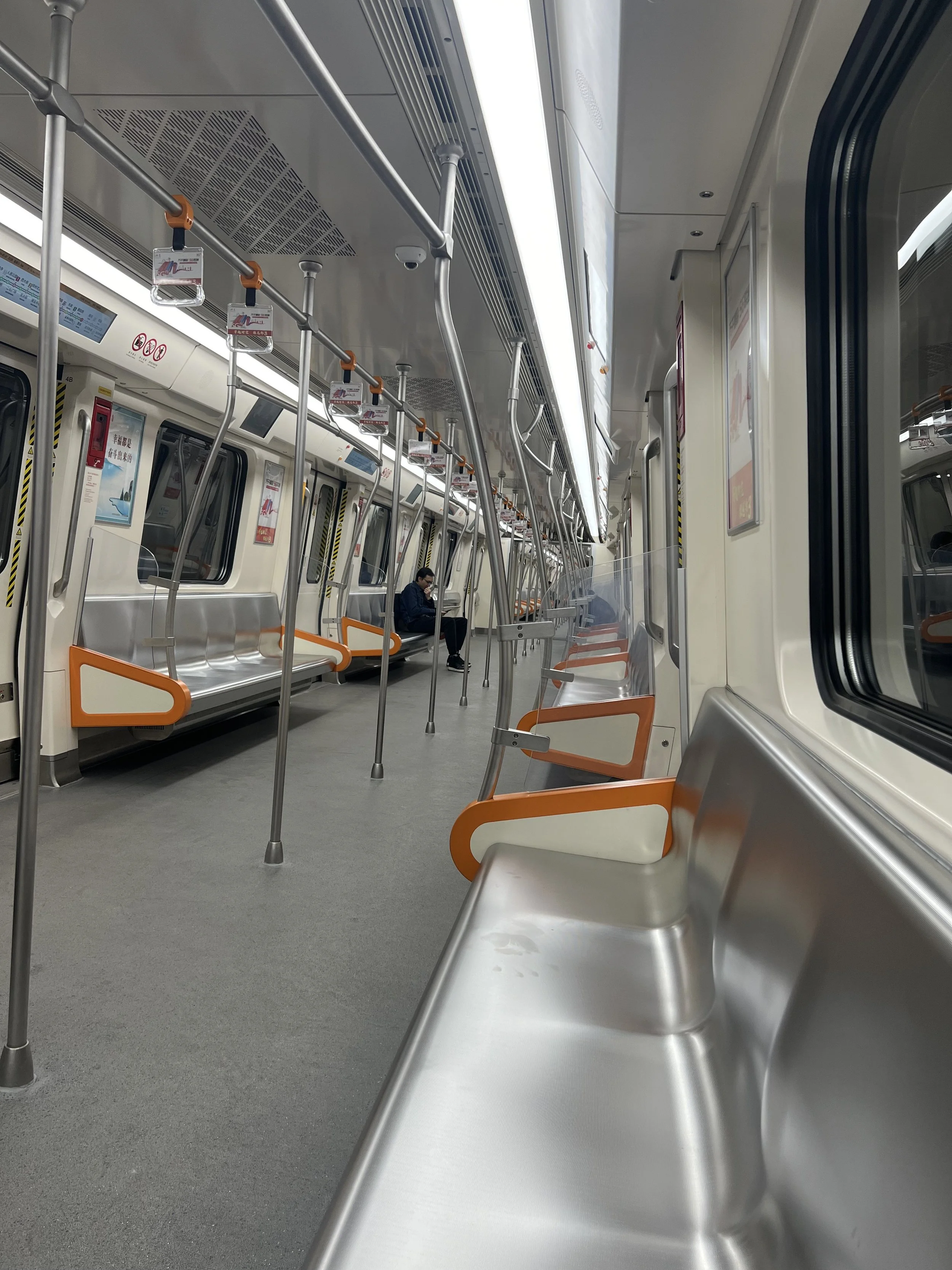Empty train car interior with a single passenger sitting alone, metal seating with orange accents, and handrails.