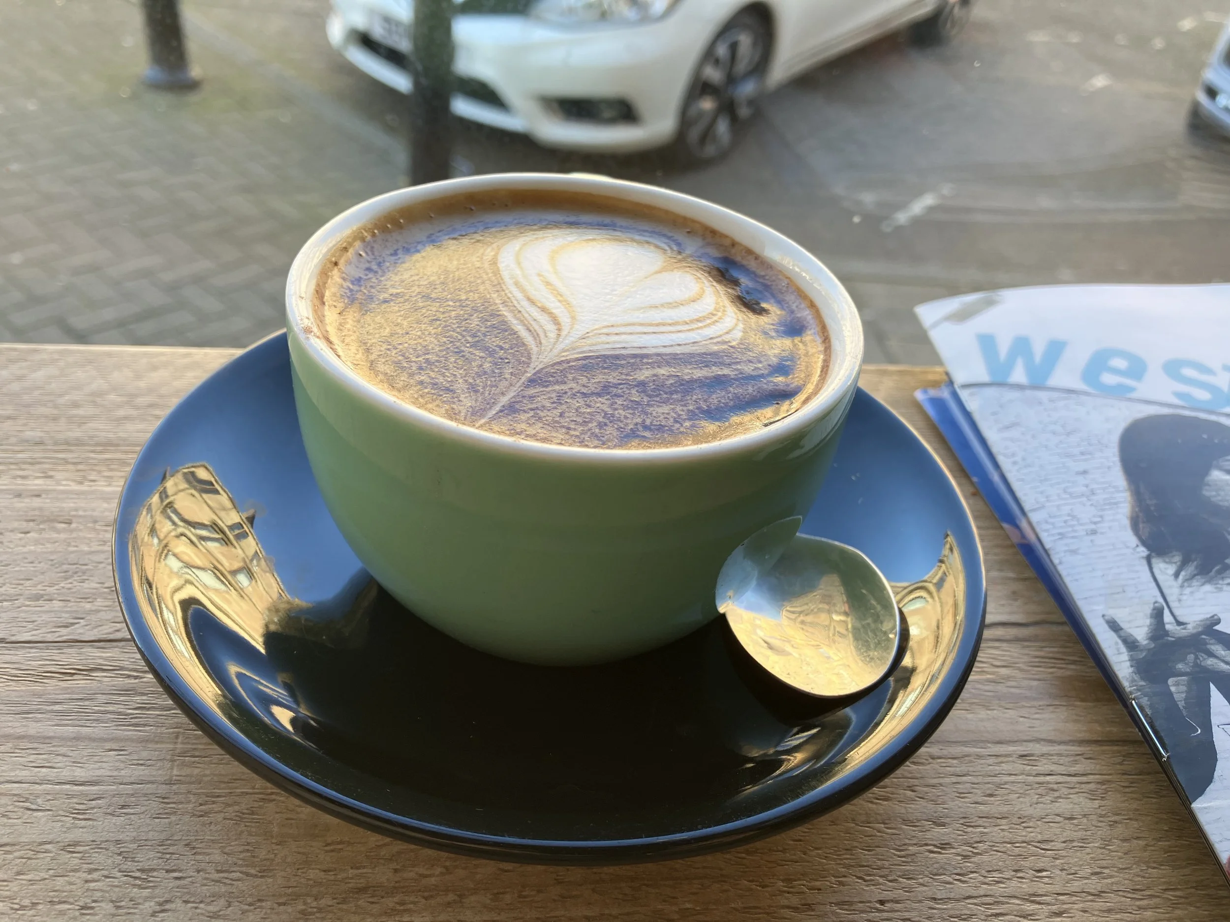 A cup of coffee with latte art in a green mug on a blue saucer, placed on a wooden table near a window with blurred cars outside. A spoon rests on the saucer and a magazine is partially visible to the right.