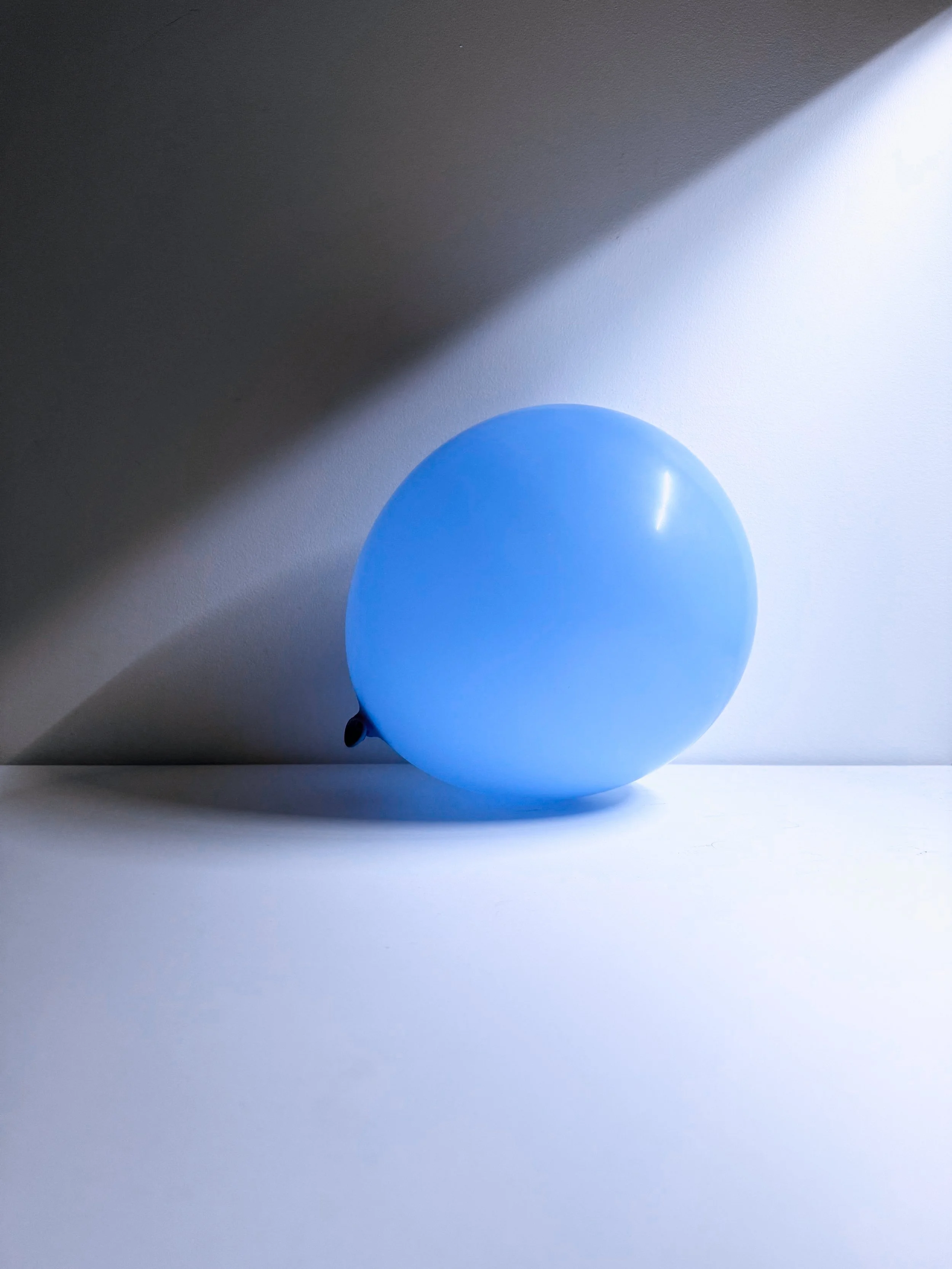 A blue balloon positioned on a white surface with a shadow cast from the top left, creating a diagonal shading pattern.