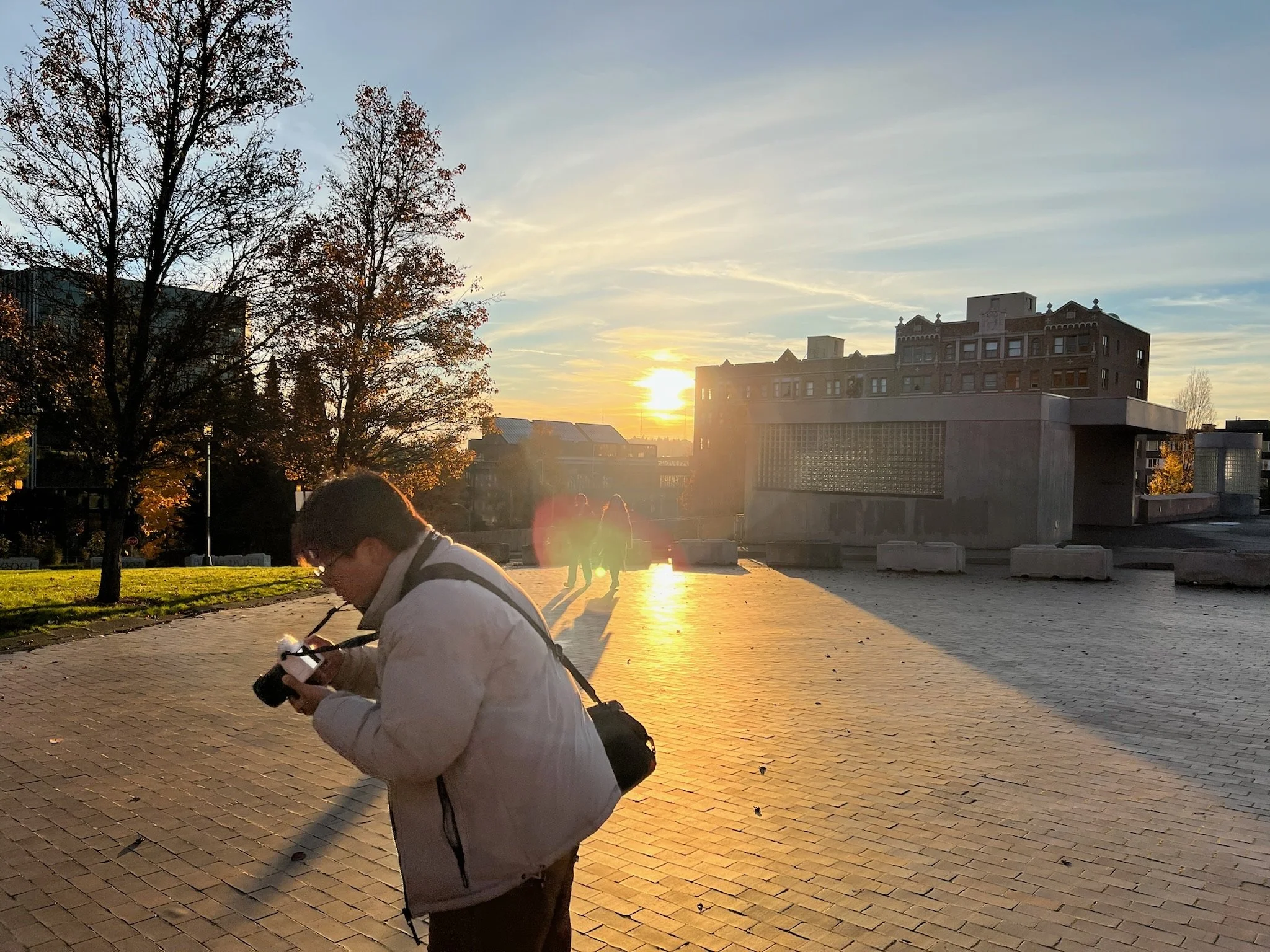 A person with glasses and a white jacket, holding a camera, is looking down at their camera while standing on a brick-paved area during sunset. Behind them, a grassy area with trees and a building with a modern architectural style can be seen, with t