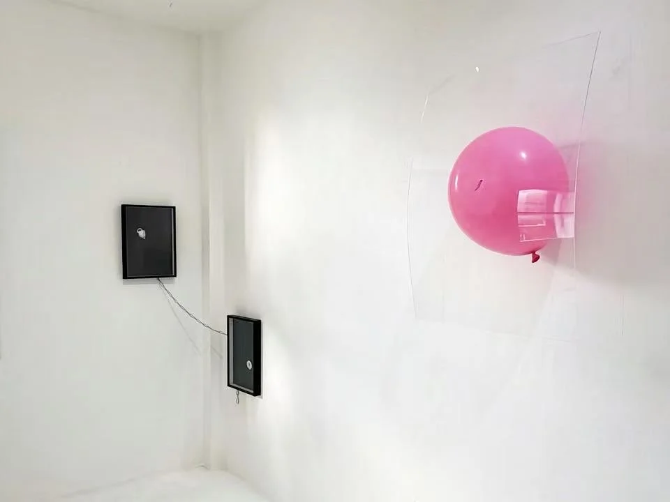 A pink balloon attached to a clear plastic sheet on a white wall with hanging black frames containing images, connected by black strings.