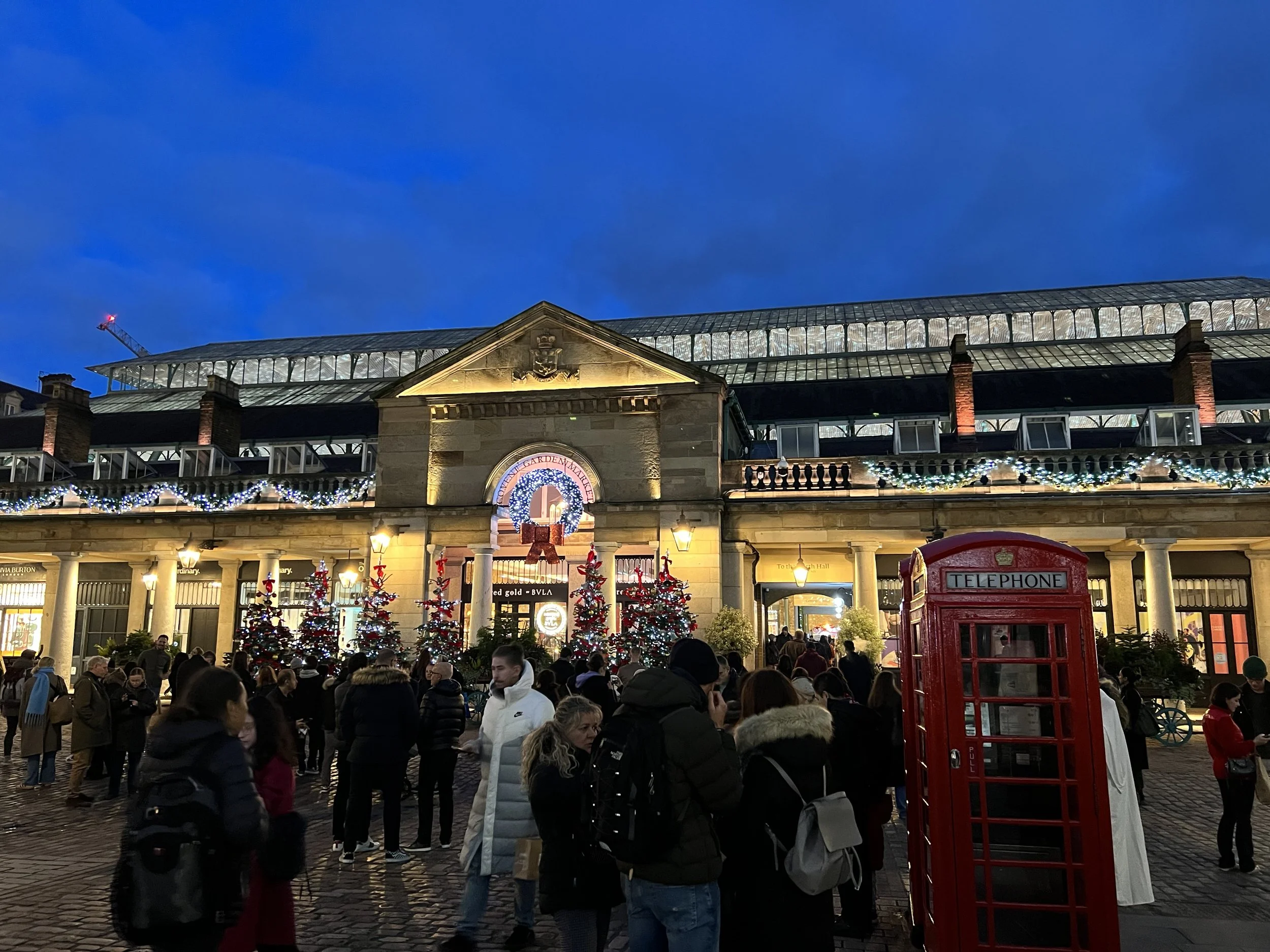 Nighttime street scene with a crowd of people gathered in front of a decorated building, including Christmas trees and festive lights. A red telephone booth is visible in the foreground.