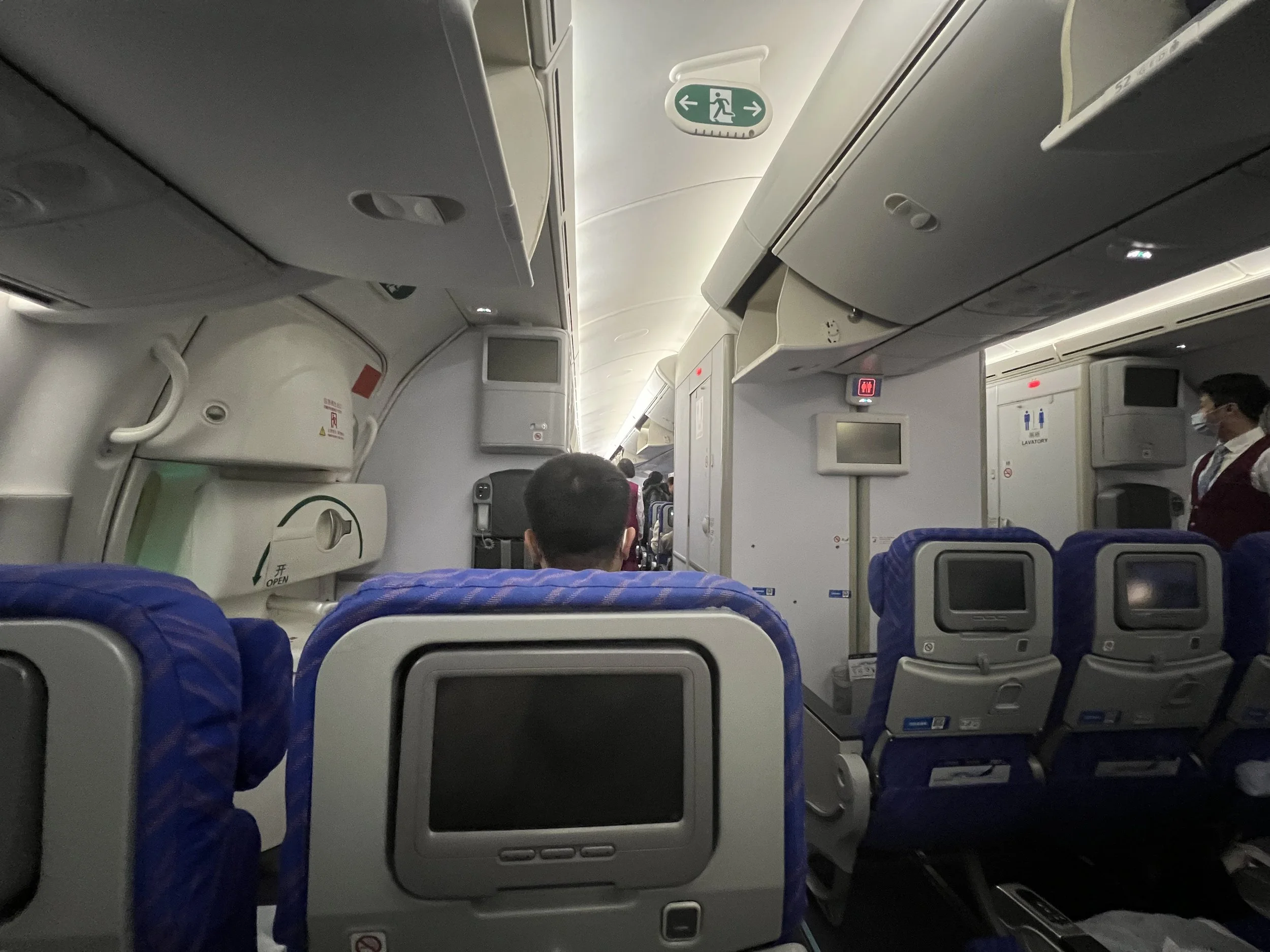 Inside view of an airplane cabin showing passenger seats, overhead compartments, a passenger wearing a face mask standing near the aisle, and emergency exit signs on the ceiling.