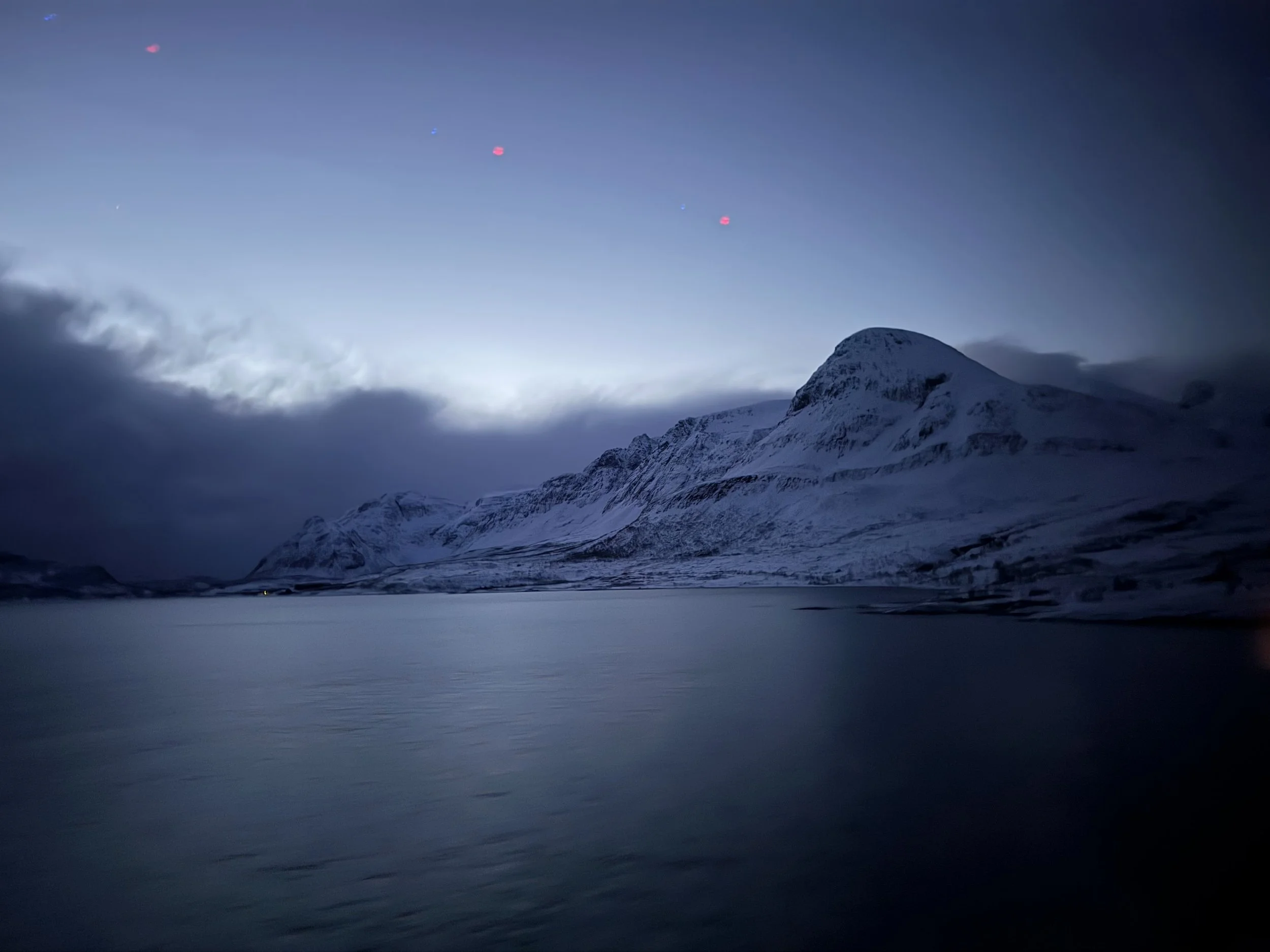 Snow-covered mountains near a calm body of water under a dark sky with some clouds and faint star or planet visible.