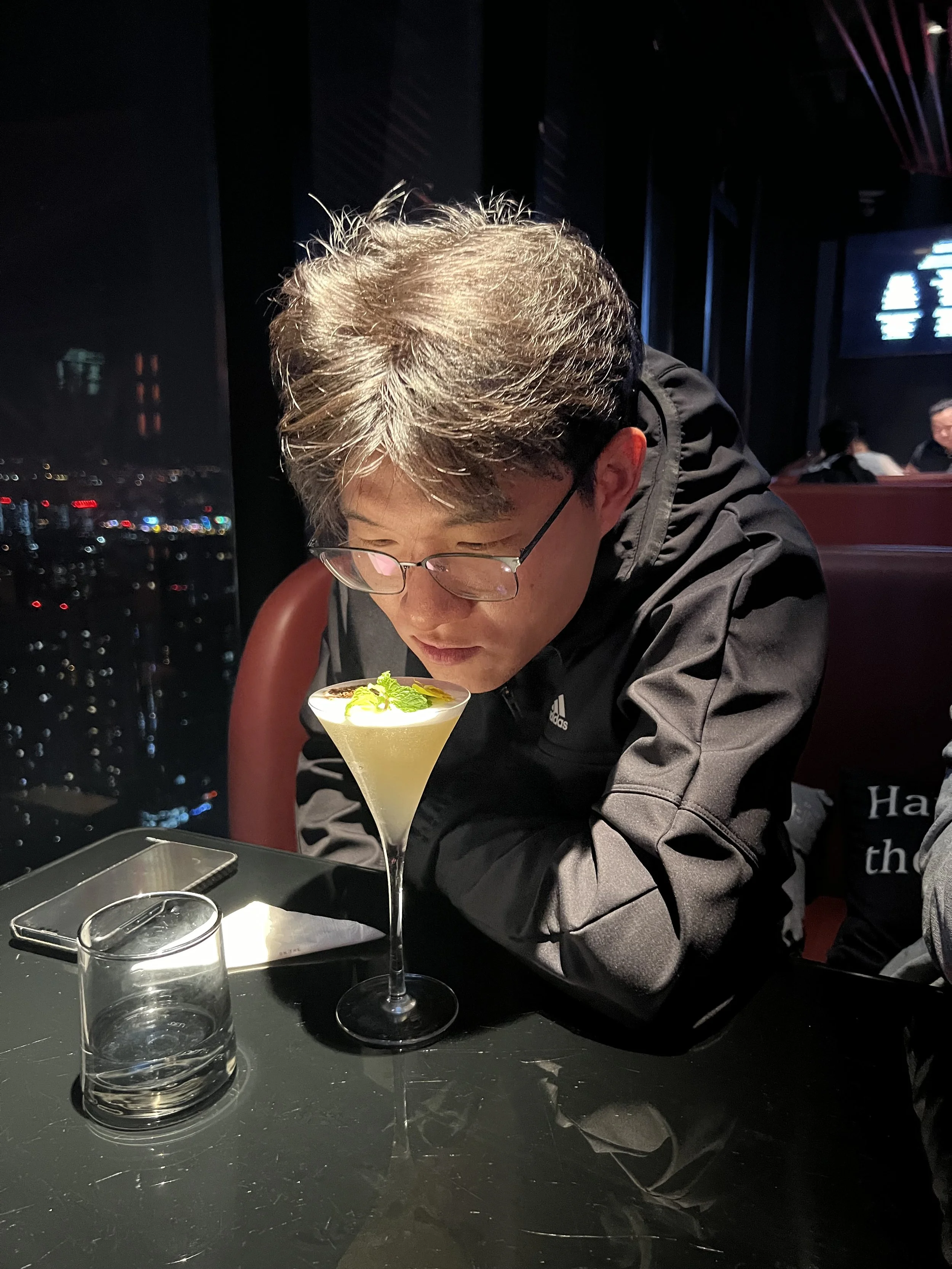 A man wearing glasses and a black Adidas jacket is sitting at a table in a dimly lit restaurant or bar, looking down at a yellow cocktail garnished with mint leaves and a lemon twist. On the table are an empty glass, a smartphone, and a small contain
