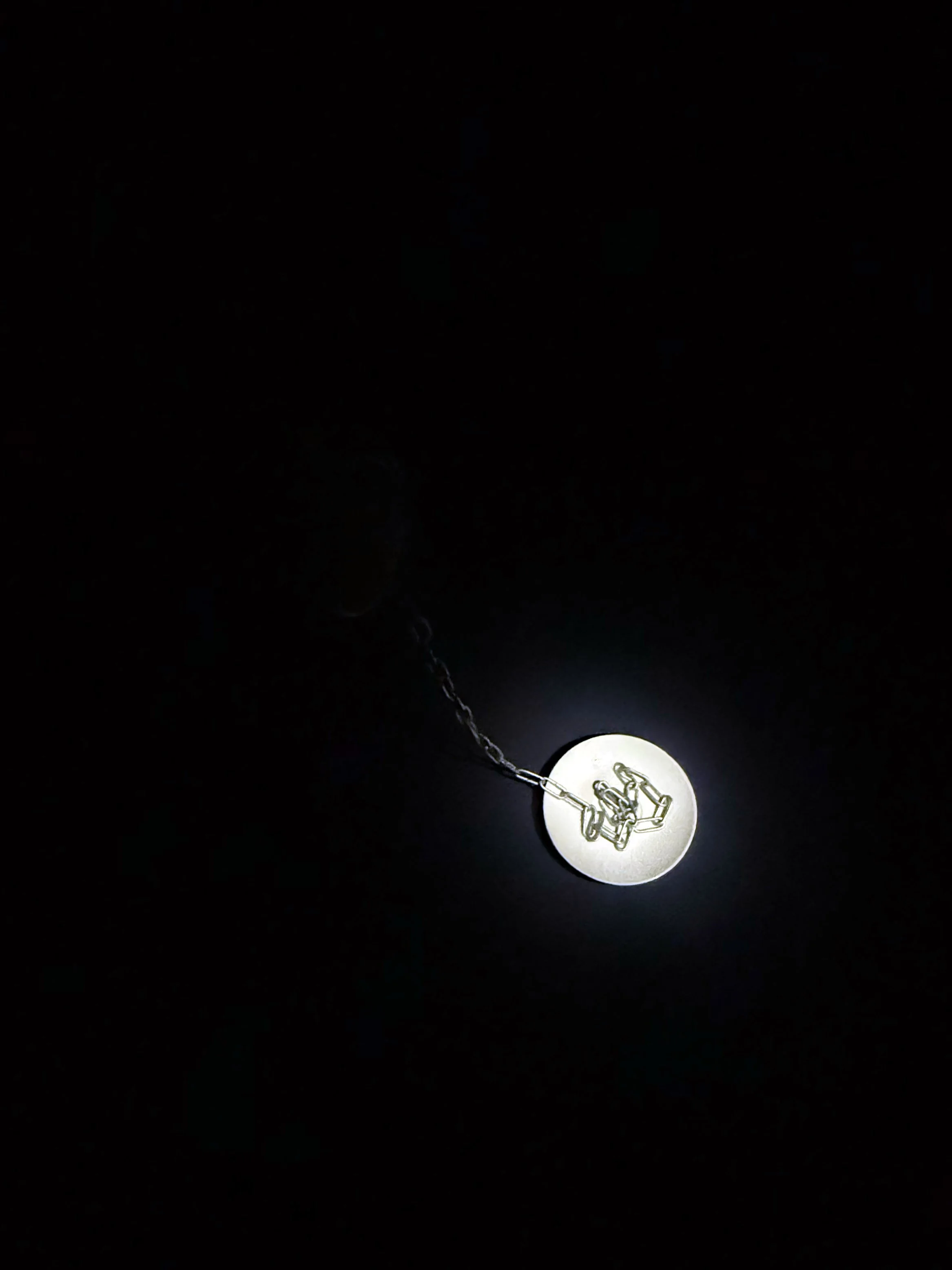 A close-up of a white circular pendant with a small, detailed metallic figure inside, hanging from a chain against a dark background.