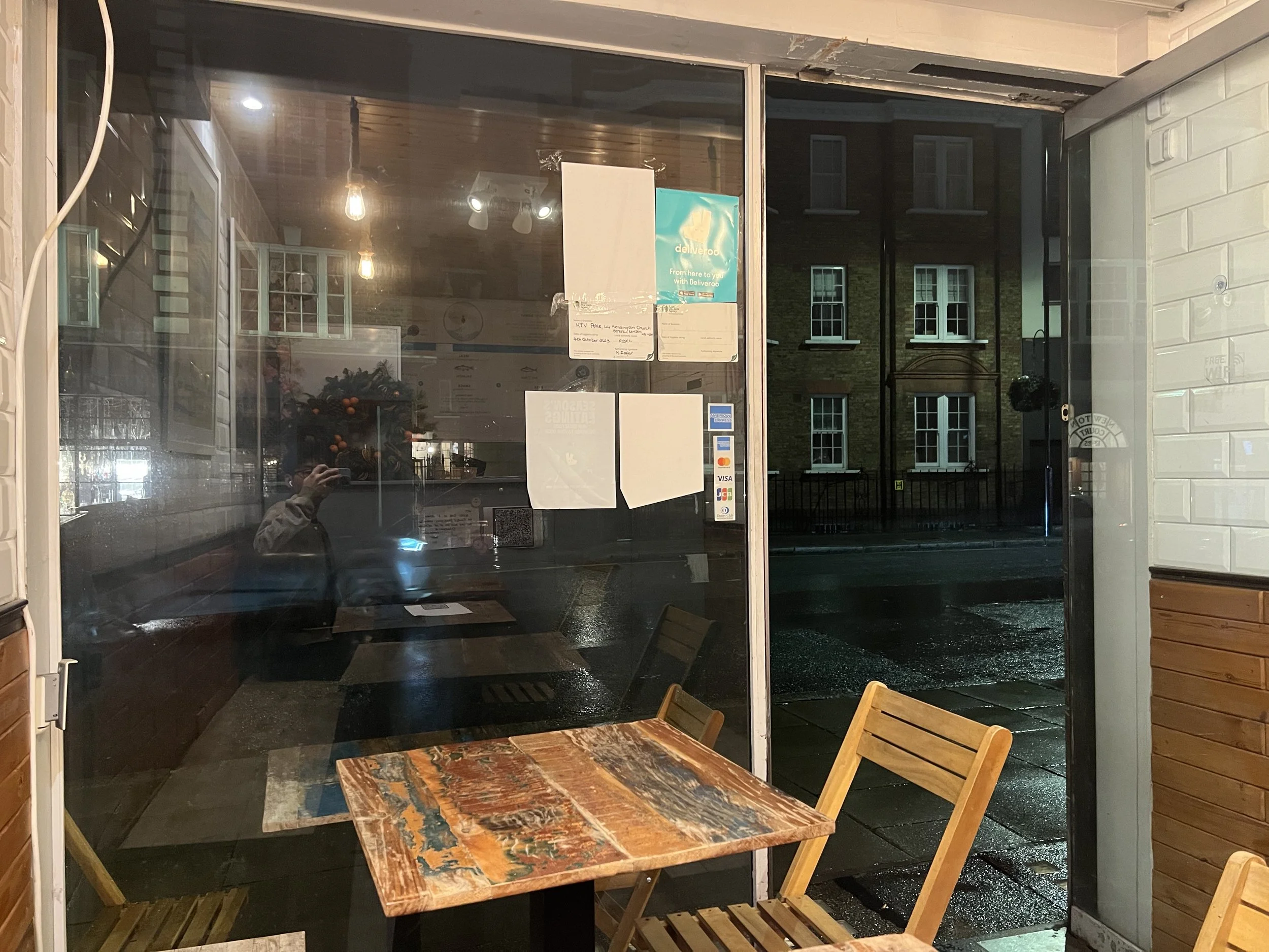 View through a glass door at a cozy indoor seating area with a wooden table and chairs. Outside, a building with multiple windows is visible during nighttime, and the scene appears wet, possibly from rain.