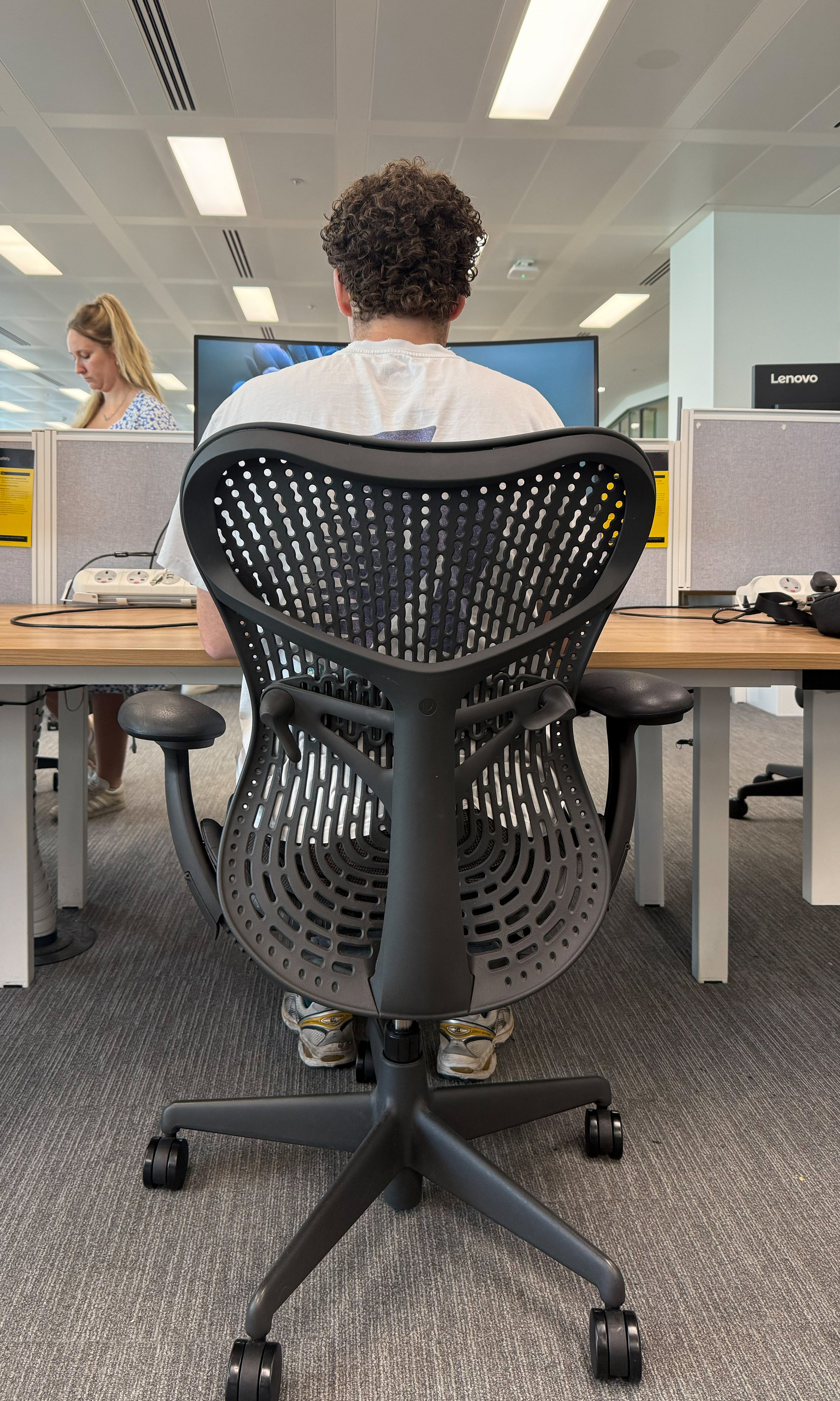 Person with curly hair sitting in an ergonomic office chair at a desk in an open-plan office, facing a monitor, with another woman working nearby.