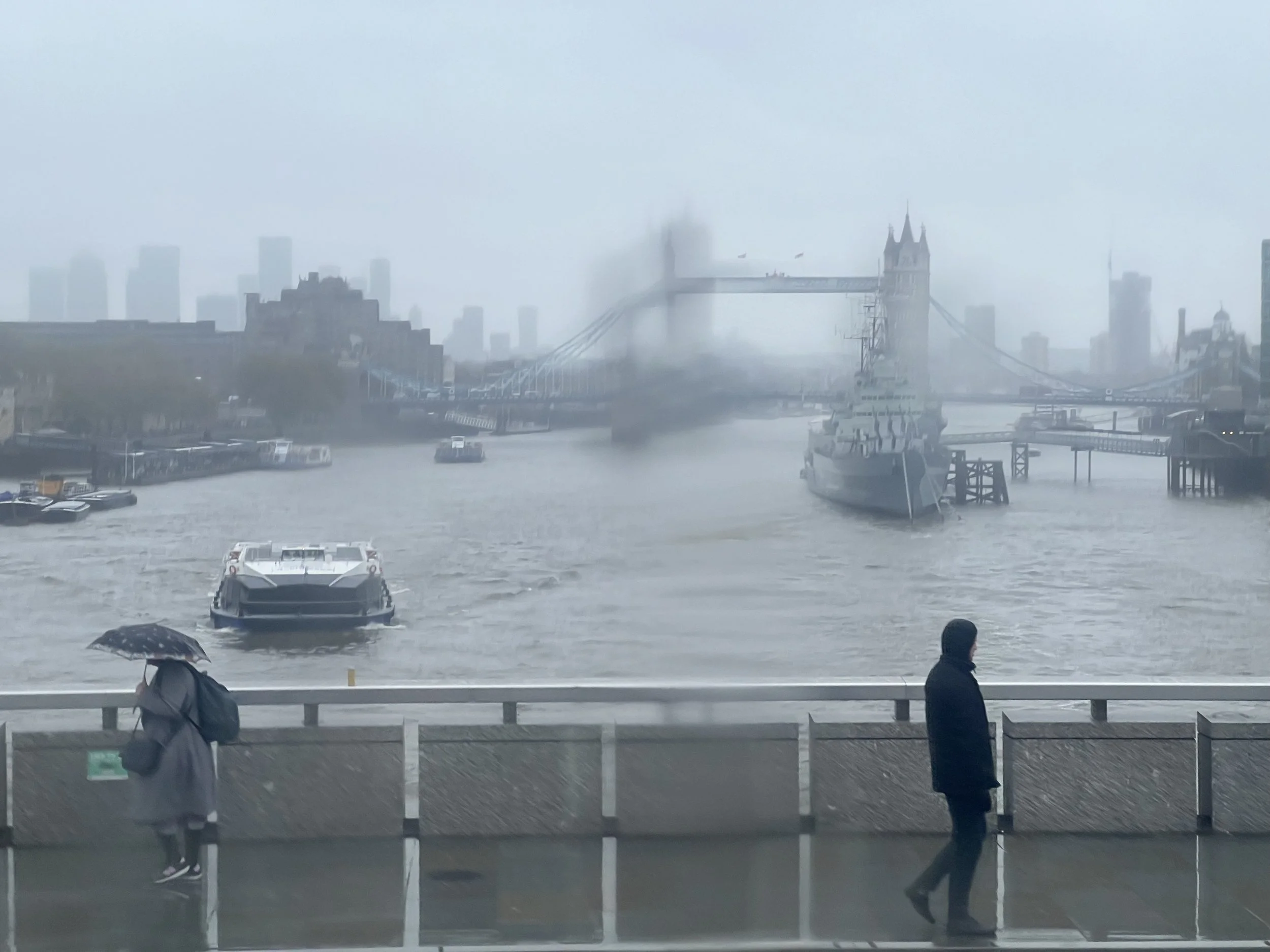 A view of the Tower Bridge over the River Thames in London during heavy rain and fog, with boats on the water and pedestrians with umbrellas on the walkway.