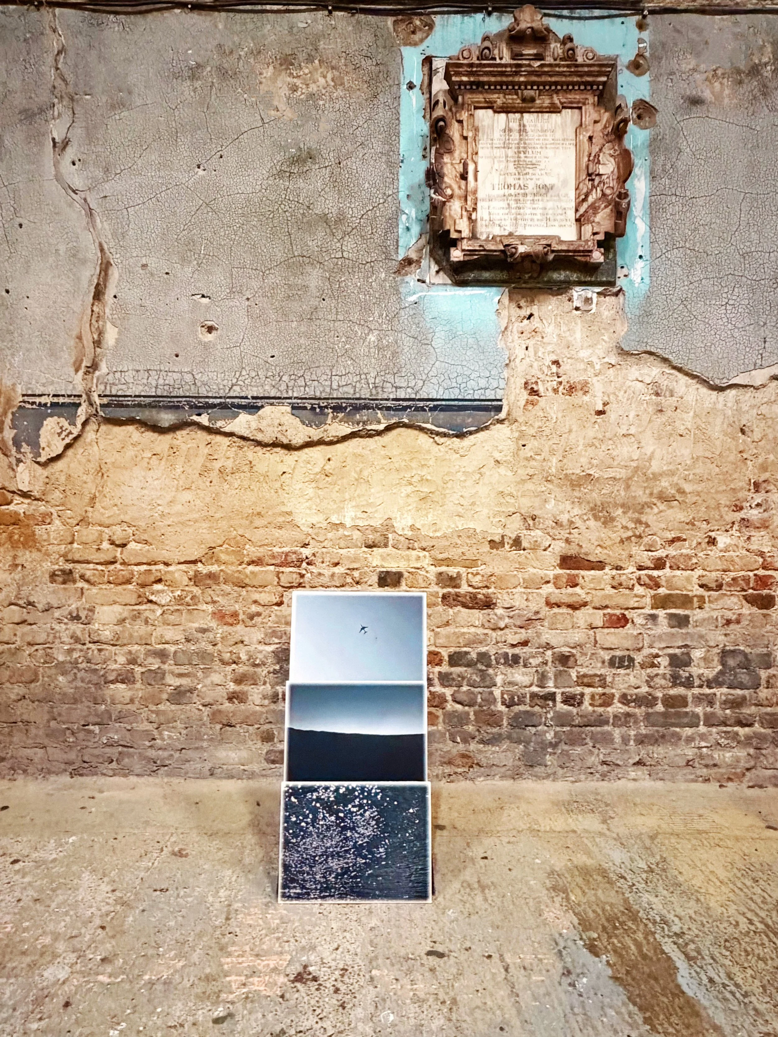 The image features a rough, cracked brick wall with an ornate, vintage wooden frame and plaque on the top right, along with a paper or card with a photograph of a plane at the bottom.