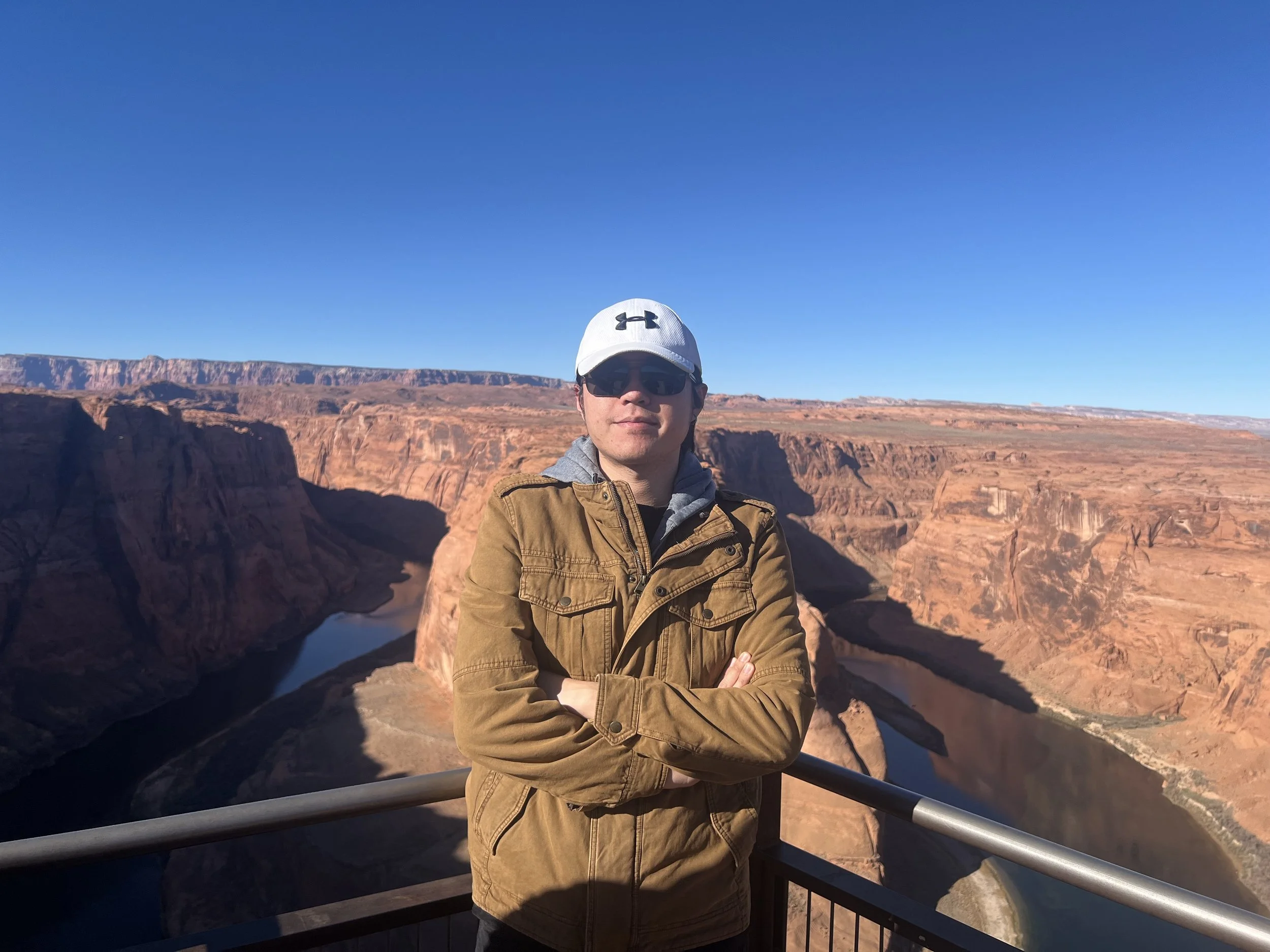 A man wearing sunglasses, a white cap with the Under Armour logo, and a brown jacket stands with arms crossed in front of a canyon. The background features red rock formations and a clear blue sky.