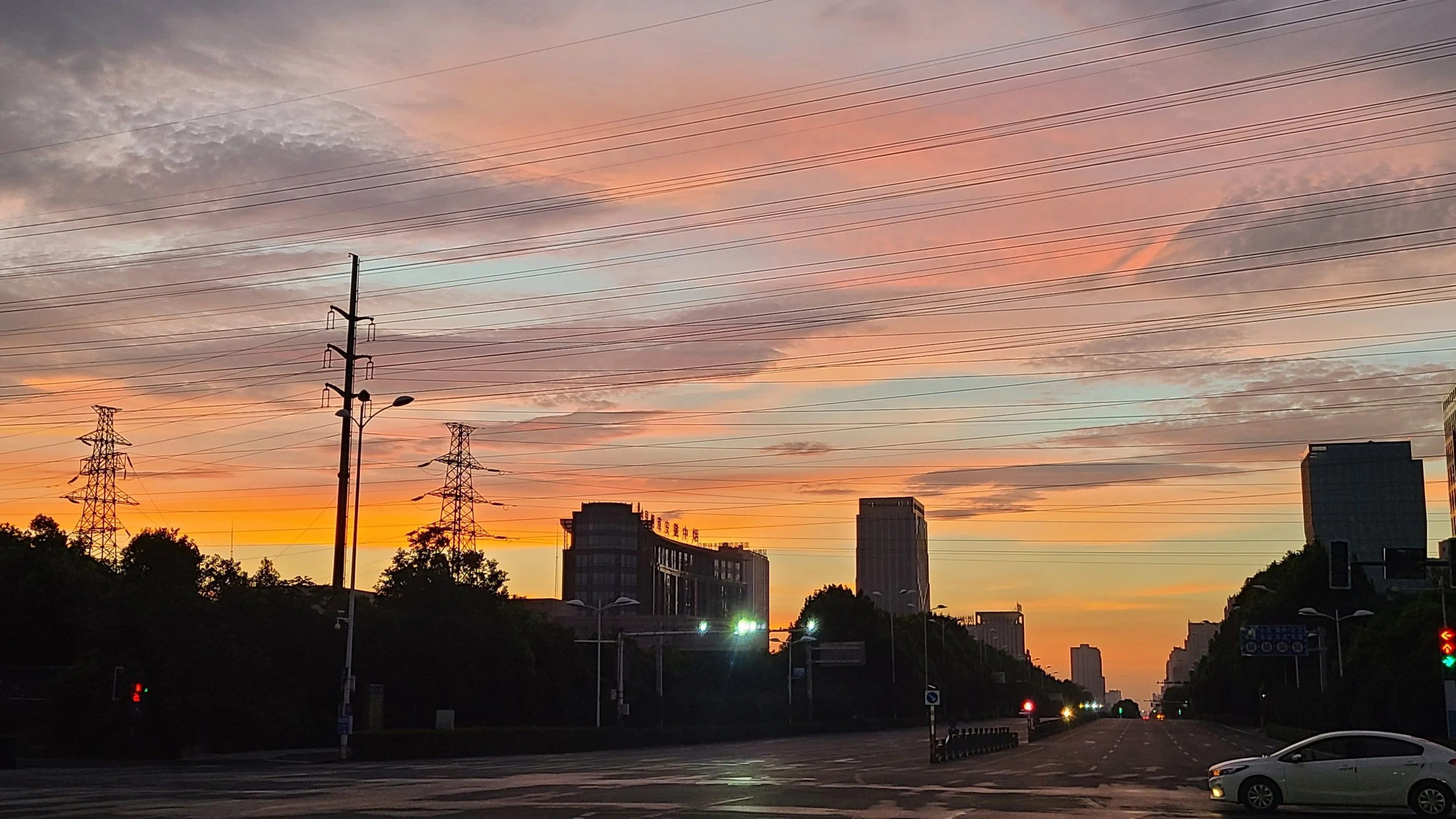 An urban sunset sky with pink, orange, and purple hues, over buildings, trees, power lines, and a street with cars.