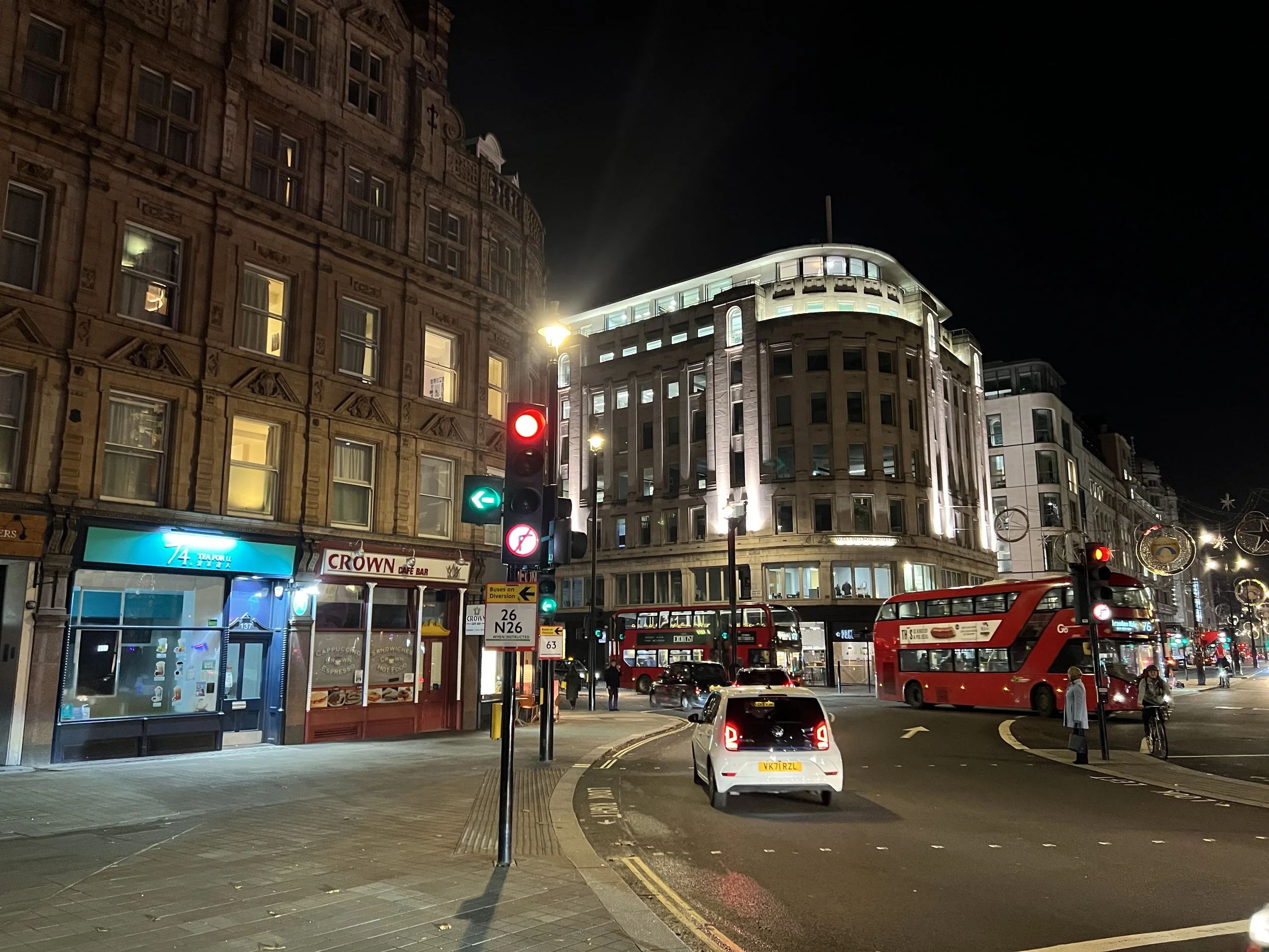 A city street at night with historical and modern buildings, red double-decker buses, cars, traffic lights, and pedestrians.