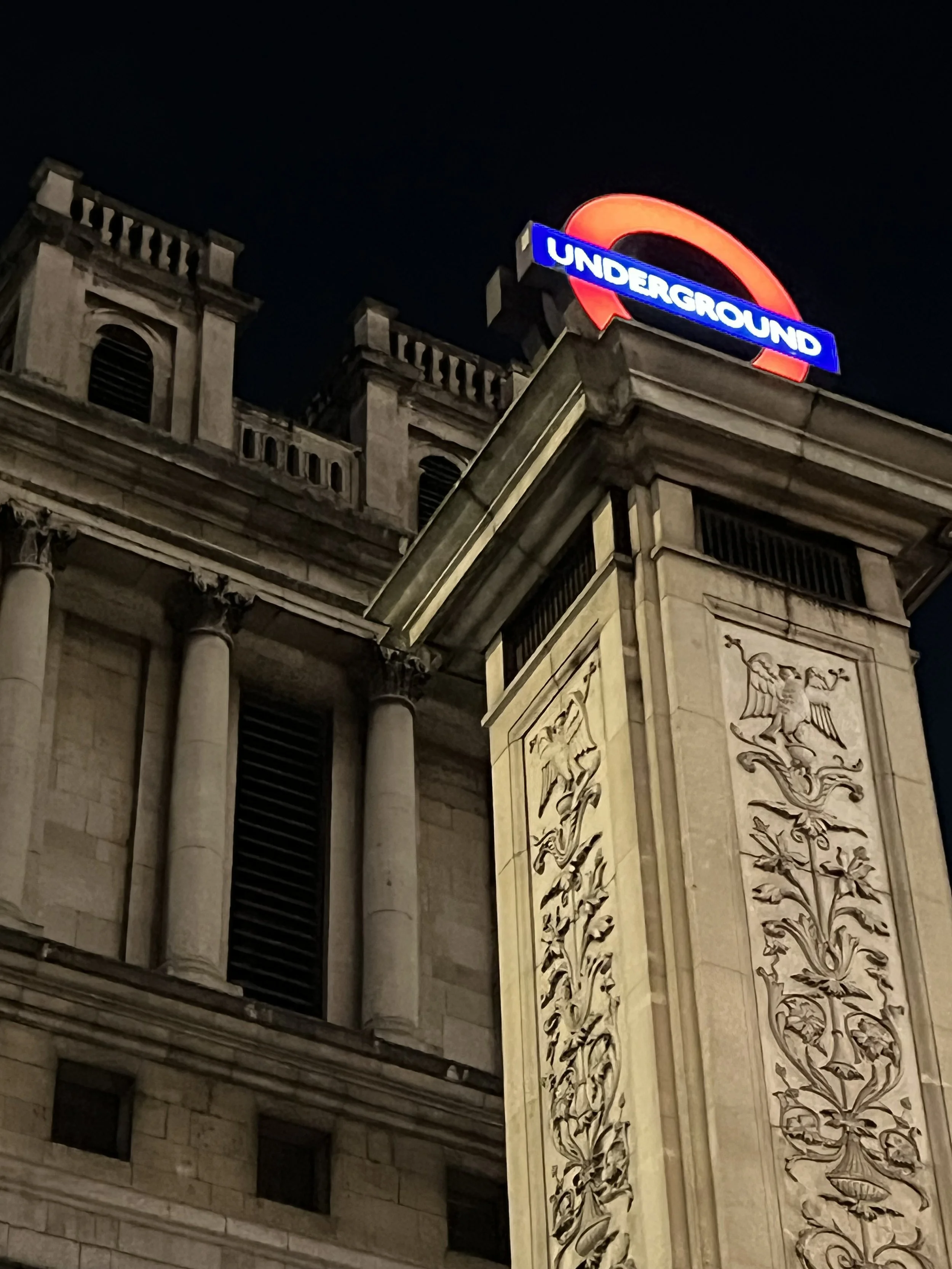 Nighttime view of a historic building with a sign reading 'Underground' in neon lights, mounted on a decorative stone wall with sculpted patterns and crests.