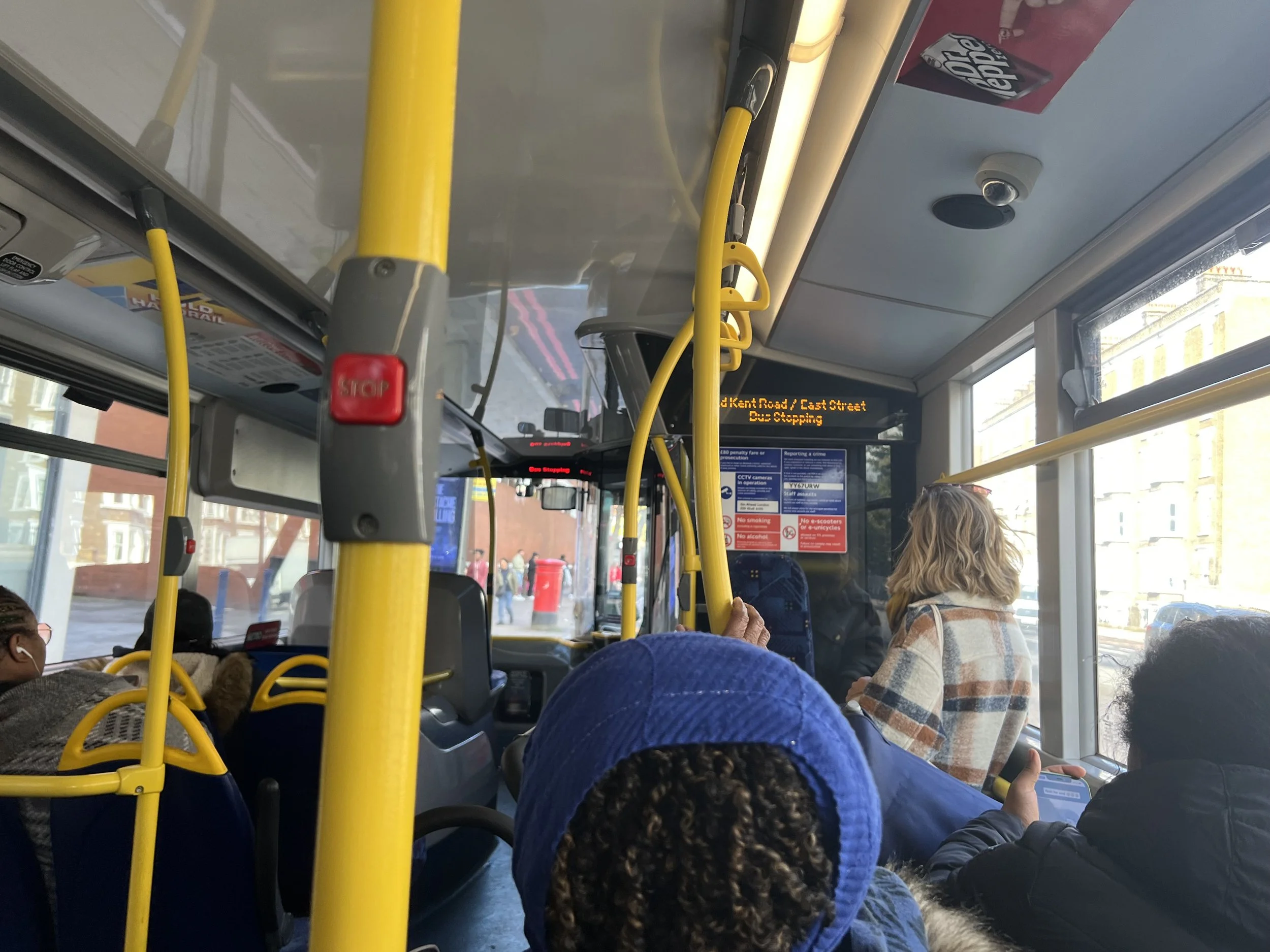 Inside of a city bus with passengers seated and standing, yellow handrails, a digital display with bus route information, and a view of buildings outside the windows.