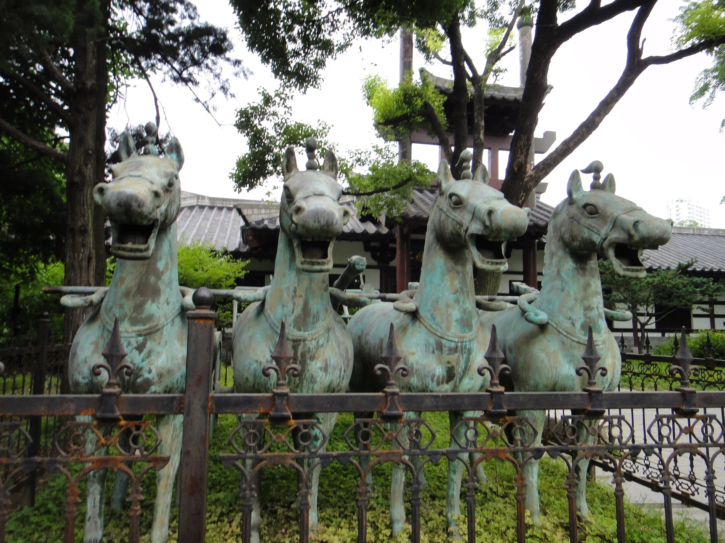Bronze statue of horses' heads, fenced and surrounded by trees in a park.
