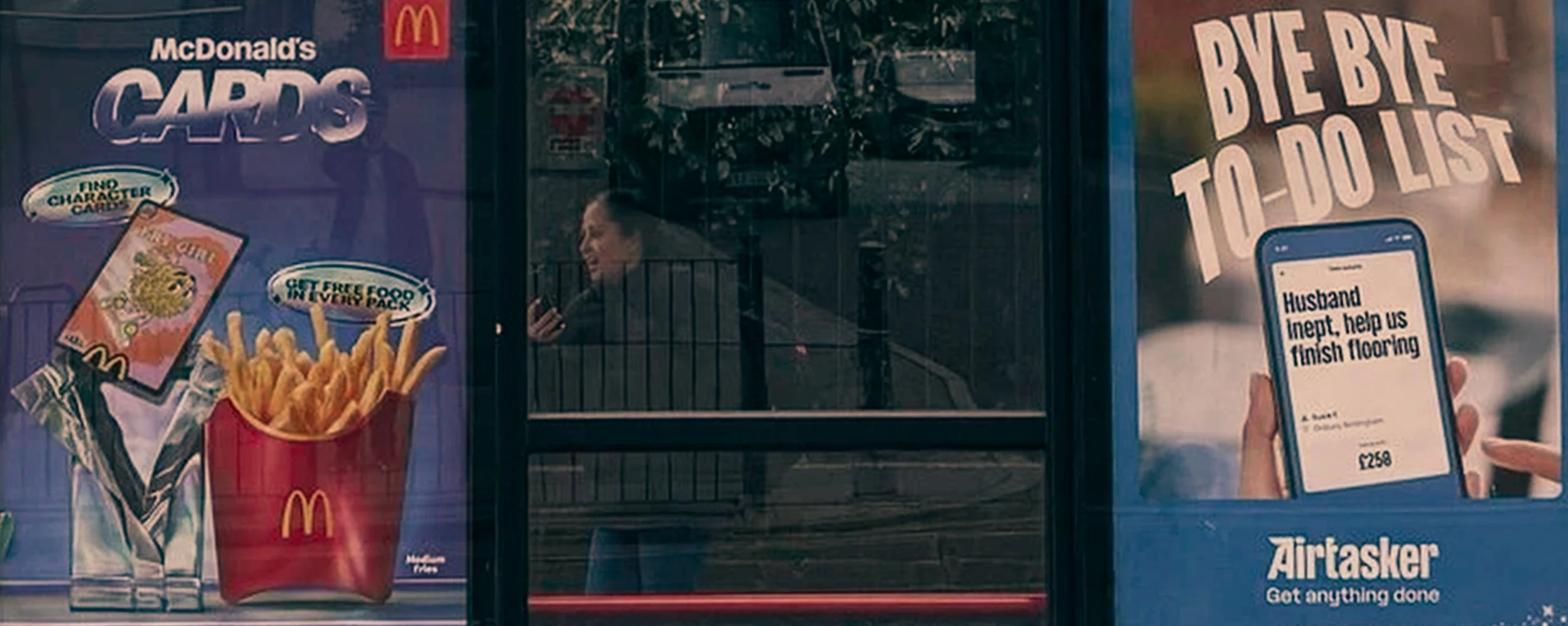 A woman inside a McDonald's restaurant looking at her phone, with advertisements for McDonald's fries and a promotion to beat the to-do list, and a sign asking for assistance to finish flooring.