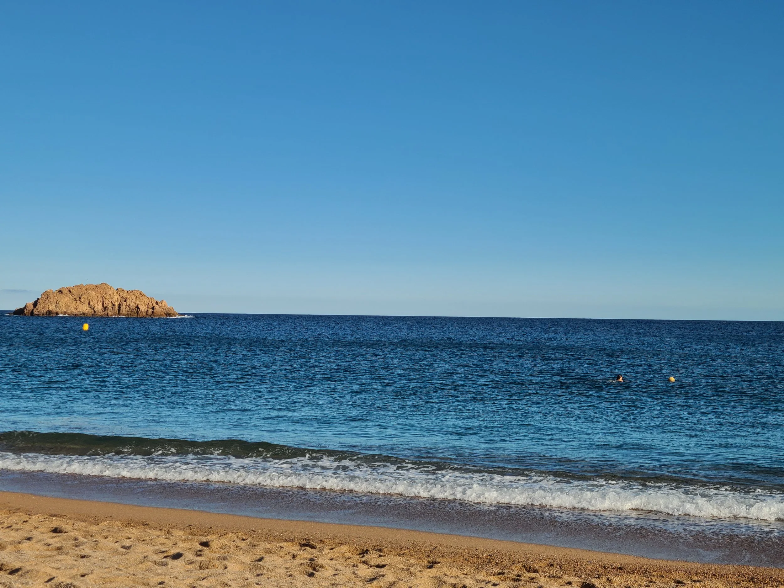 A beach scene with sand in the foreground, gentle waves, and a large rock formation offshore, under a clear blue sky.