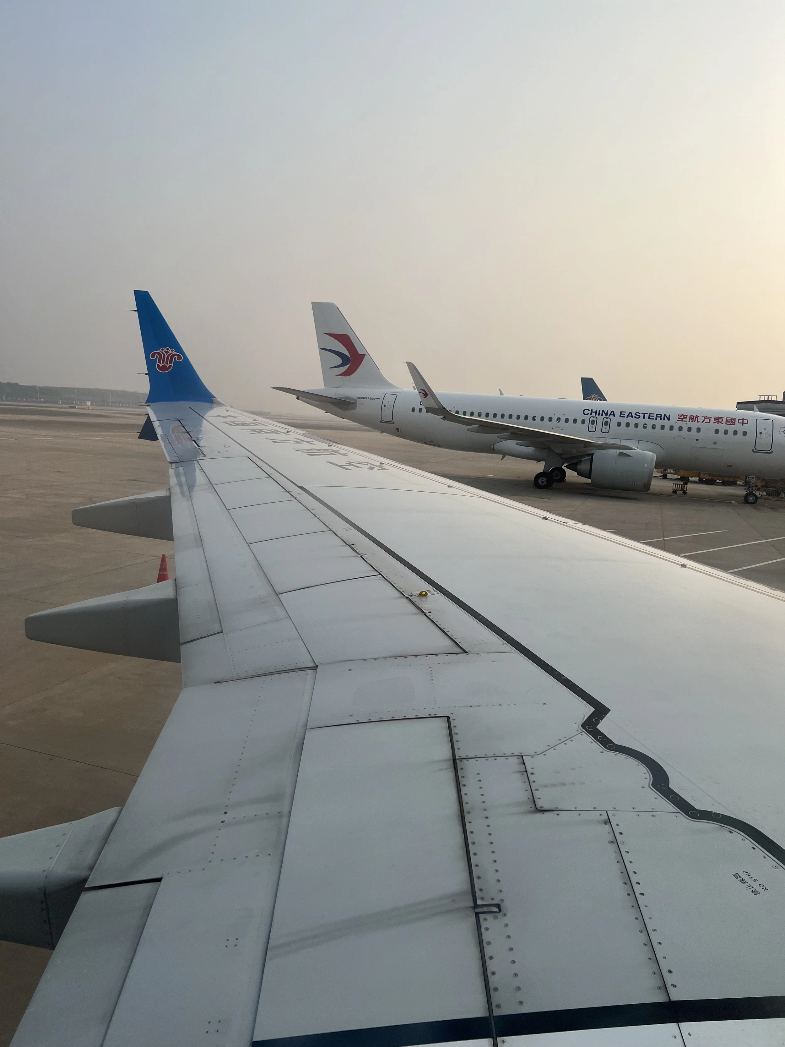 View of airplane wings and parked airplanes on the tarmac at an airport during sunset or sunrise.