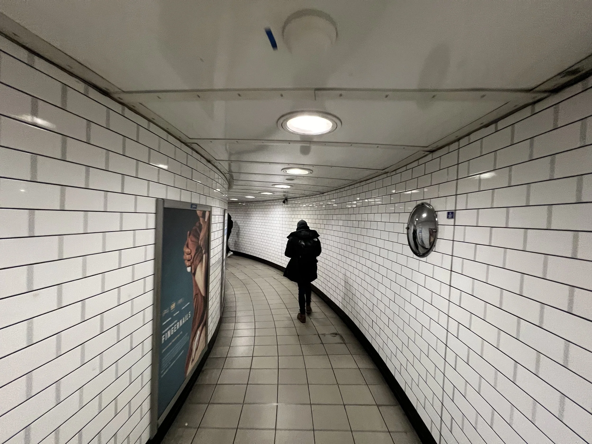 A person wearing a black coat and a hat walking down a curved underground subway corridor with tiled white walls and ceiling, illuminated by circular ceiling lights.