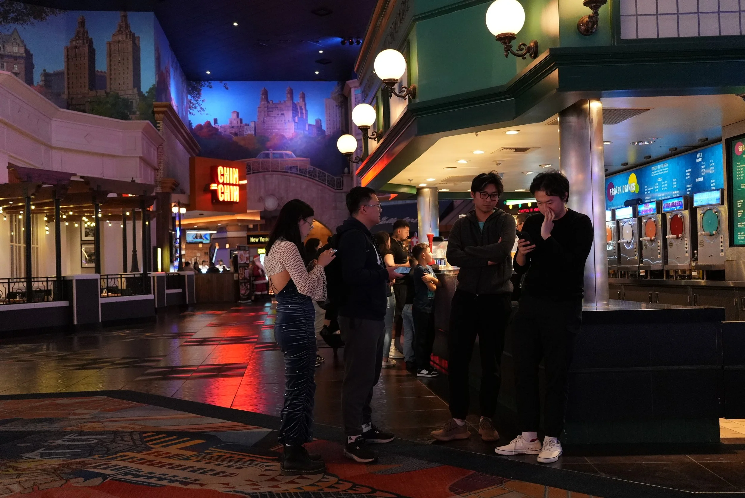 People standing in line at a food court inside a mall with mural of New York City skyline in the background, illuminated by globe lights on green walls.