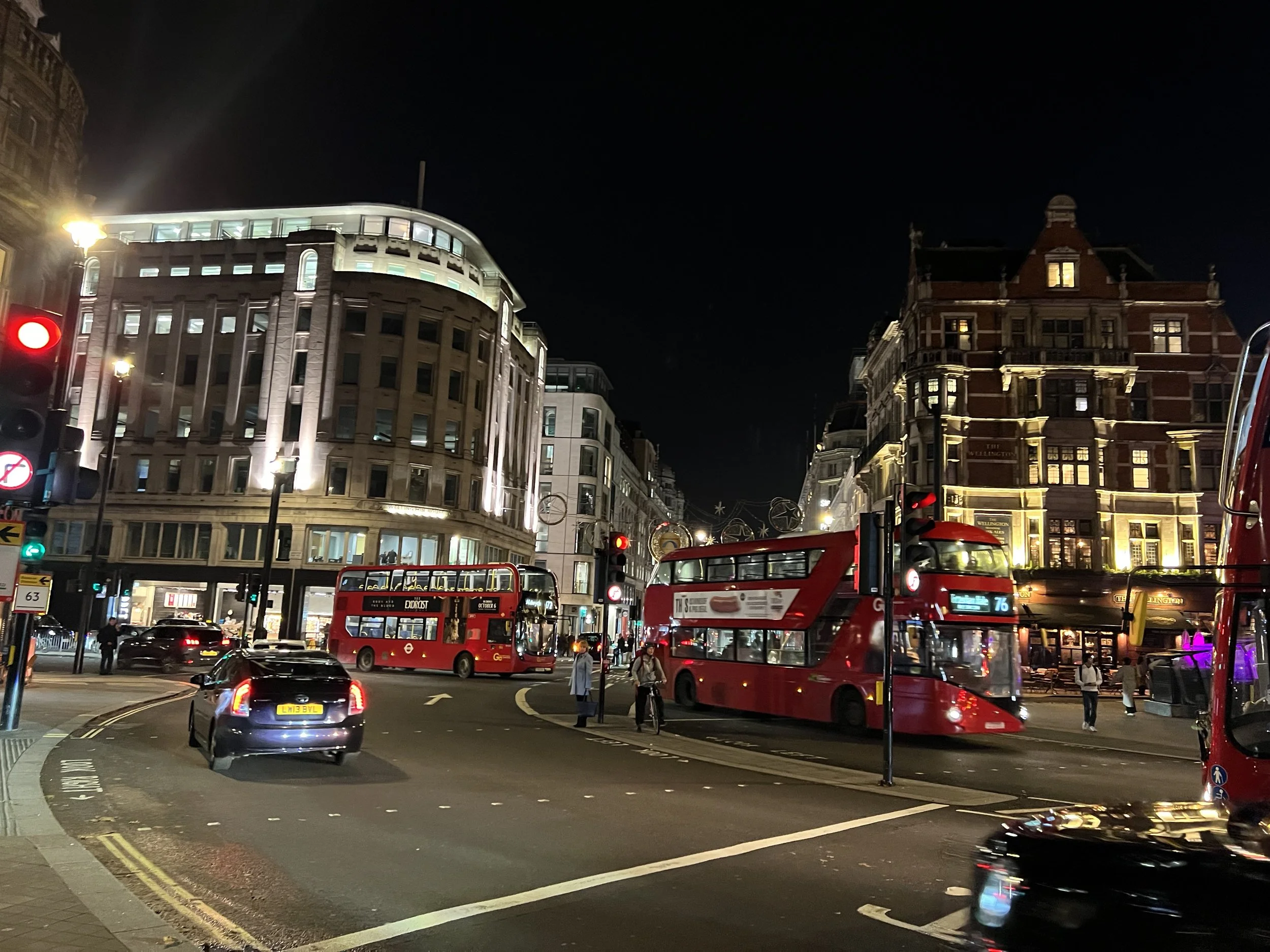 Nighttime city street scene with illuminated buildings, red double-decker buses, cars, traffic lights, and pedestrians.