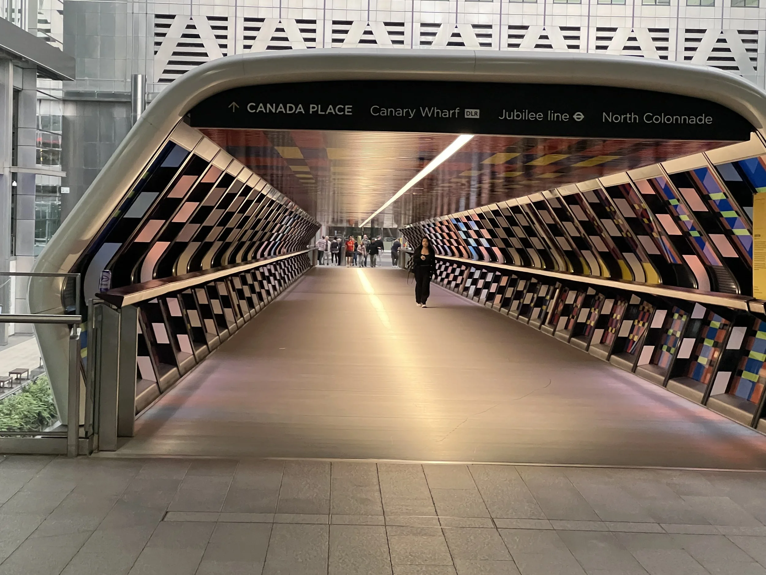 The image shows a modern, colorful pedestrian walkway or bridge inside a building, leading to an outdoor area where several people are walking. The ceiling has a sleek design with a light fixture running along the center. Above the walkway, a black d