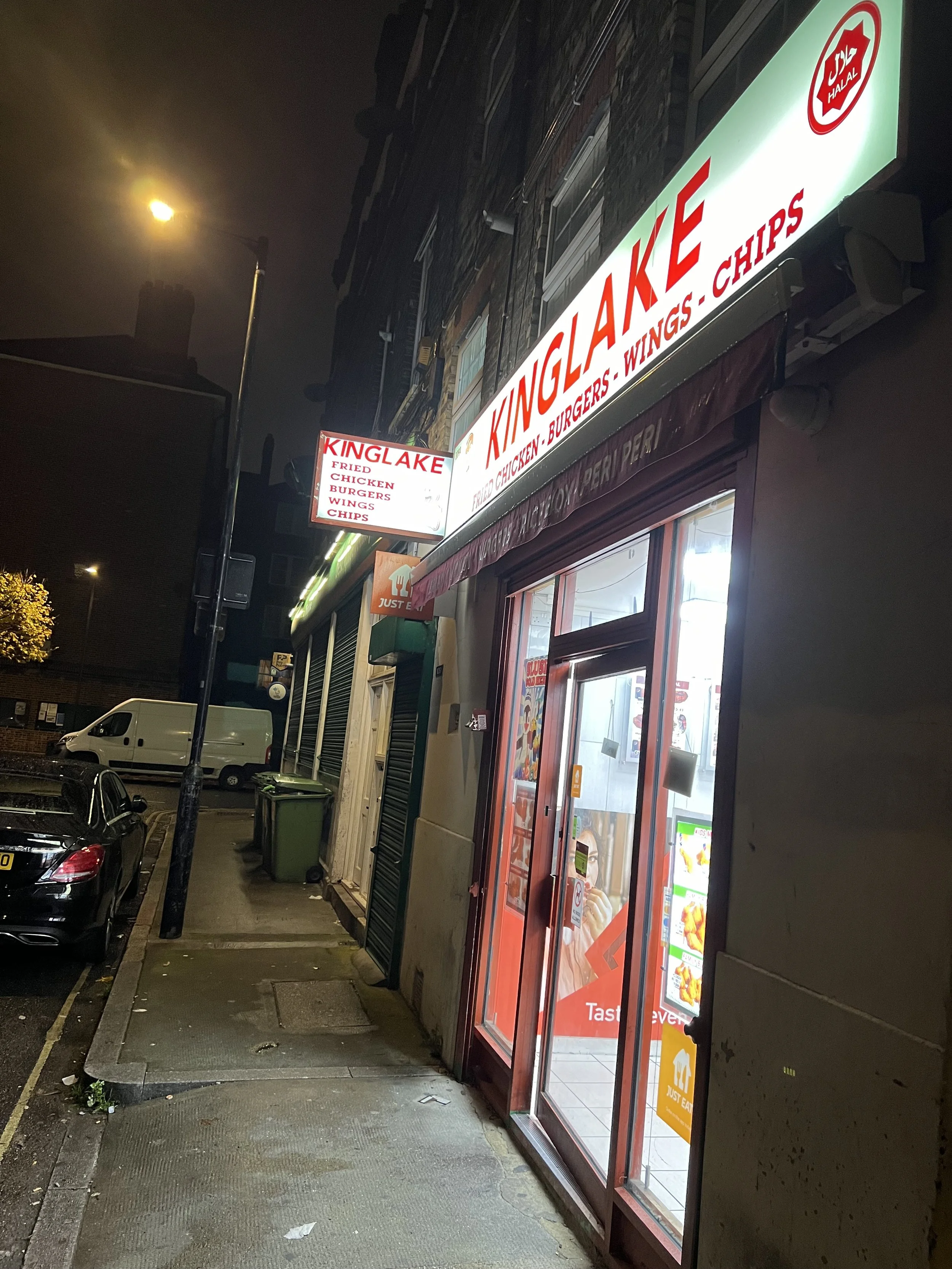Nighttime street scene showing a fast food restaurant with illuminated signs displaying menu items such as fried chicken, burgers, wings, and chips. The restaurant is located in a building with a brick facade and a large glass door. Cars are parked a