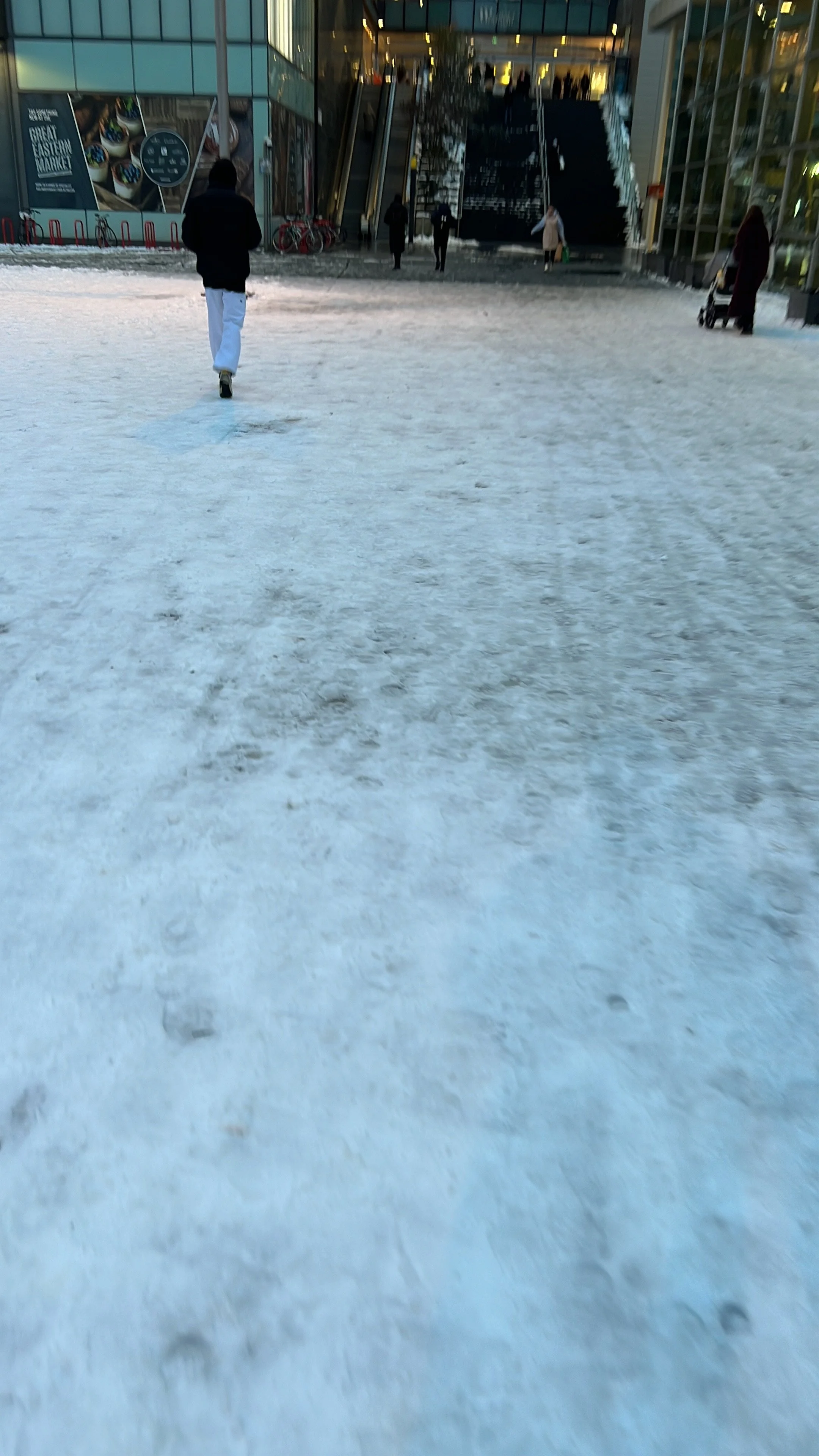 People walking outside in front of a modern building with glass walls, escalators, and a snow-covered ground.