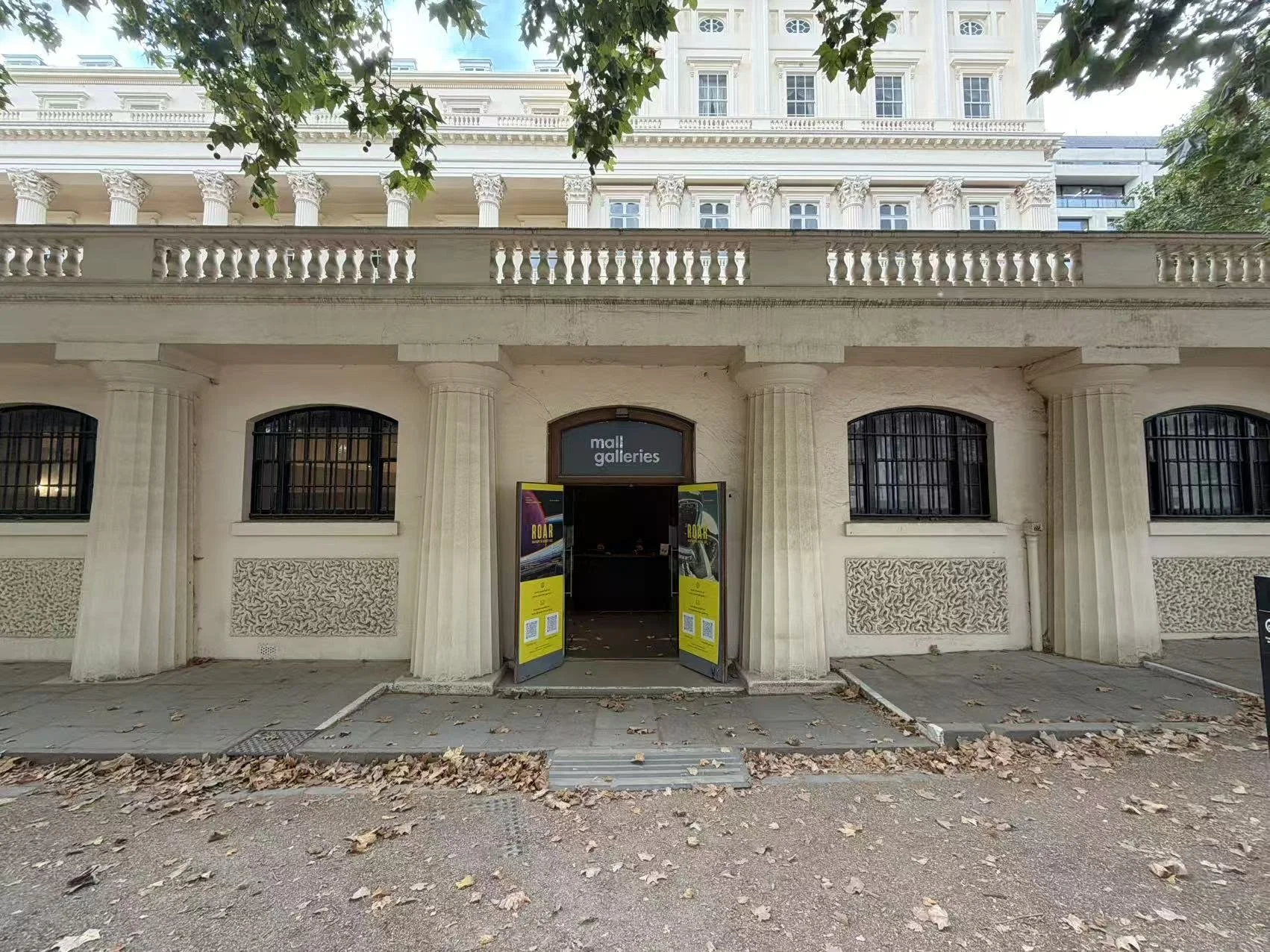 The front of a building with columns and a sign that reads 'mall galleries'. The building has barred windows, a balcony, and some fallen leaves on the ground.