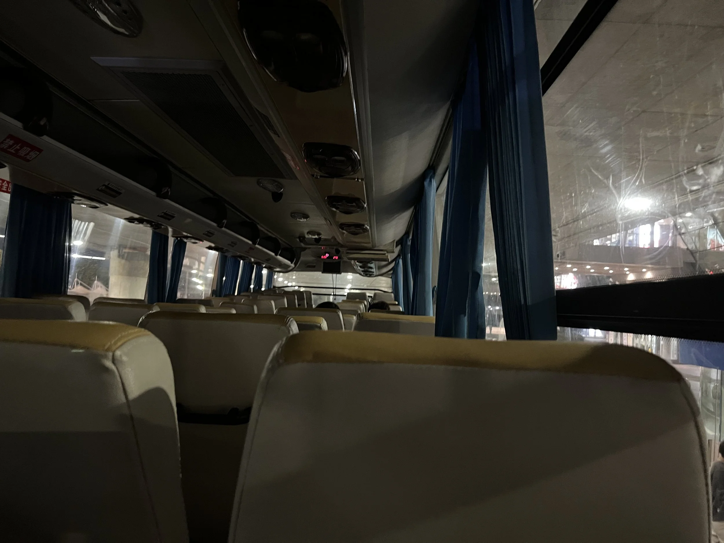 Inside view of a bus with beige seats, blue curtains, and a ceiling with air conditioning vents and lighting fixtures, in a dimly lit or overcast environment.