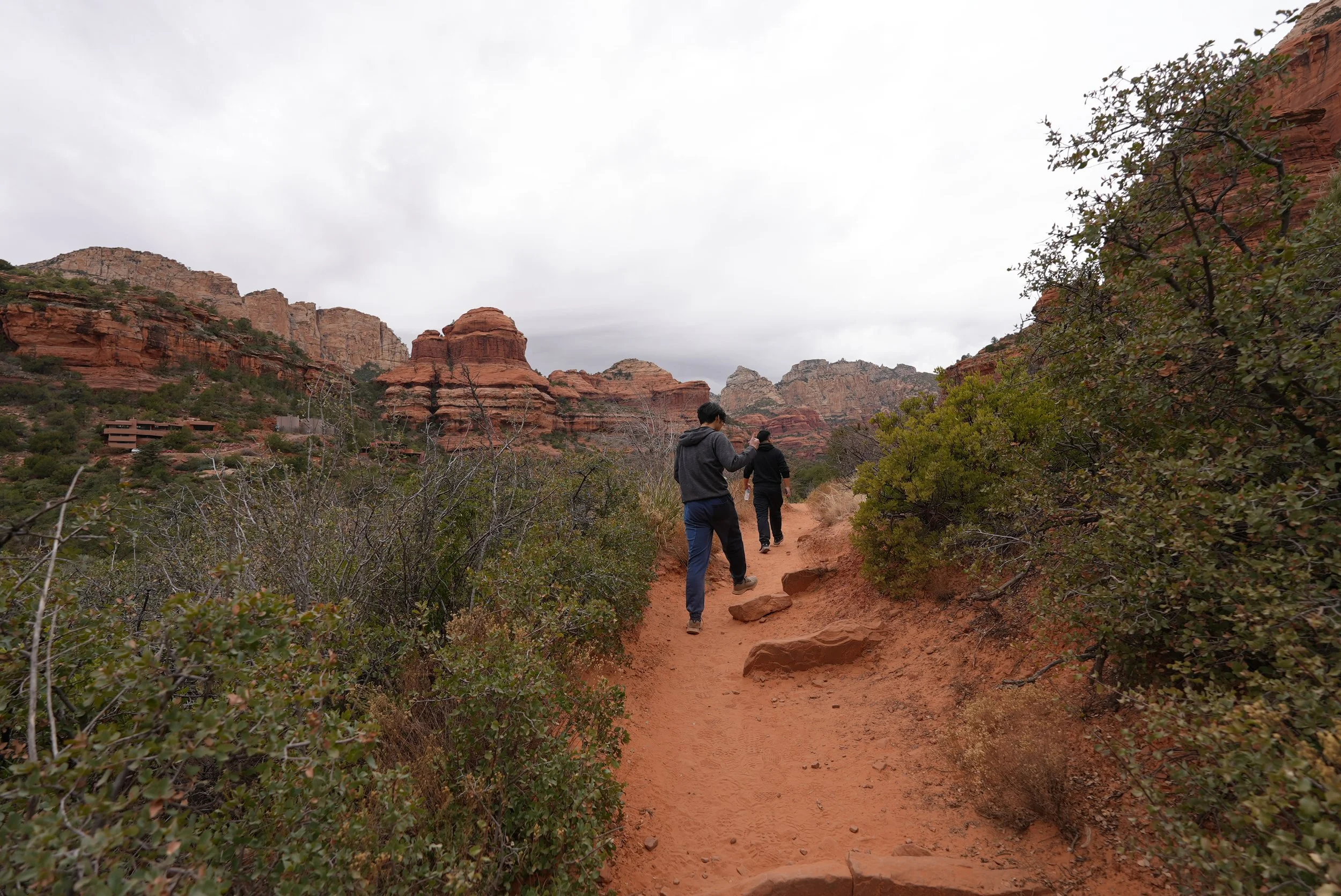 Two people walking on a dirt trail surrounded by desert shrubs and red rock formations under a cloudy sky.