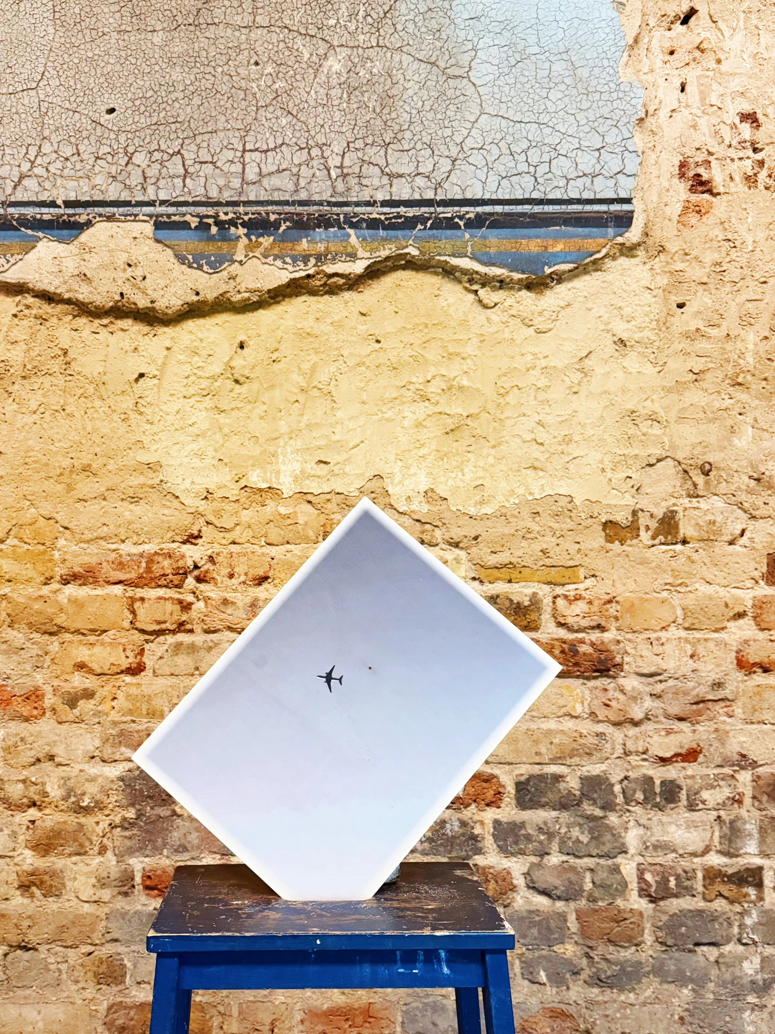 A white piece of paper with a black airplane silhouette is resting against a dark blue stool, with an exposed brick wall in the background.