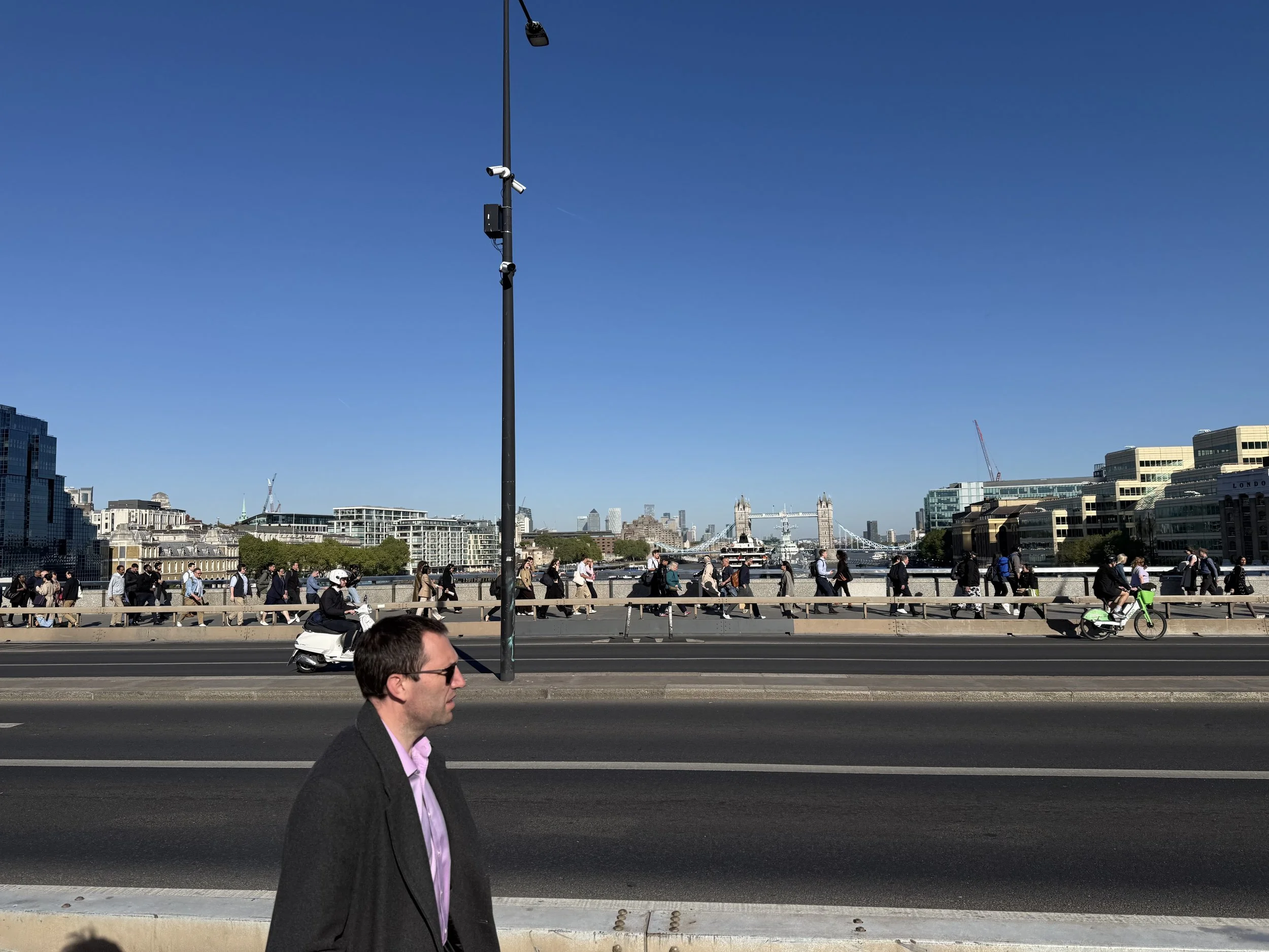 People walking and cycling along a riverbank in London with Tower Bridge in the distance, set against a clear blue sky.