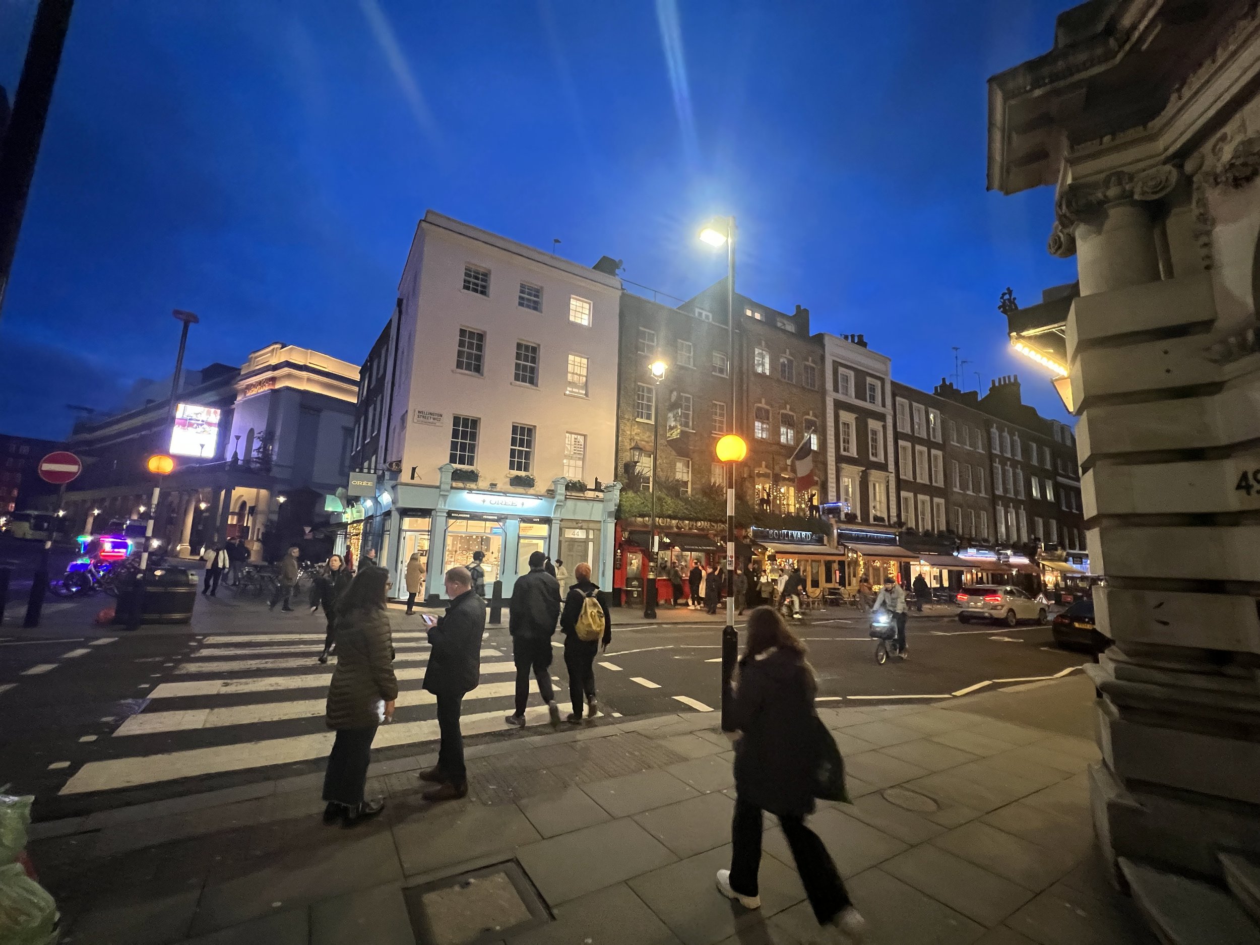 City street scene at dusk with pedestrians crossing crosswalk, buildings with lit windows, streetlights, and a clear evening sky.