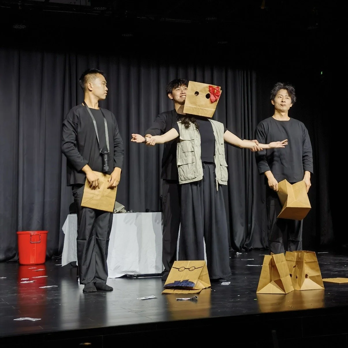 Three performers on stage in black clothing, one with a paper bag over their head featuring a red bow, acting out a scene with paper bags as props. There is a black curtain backdrop, a small table, and a red bucket on the stage.