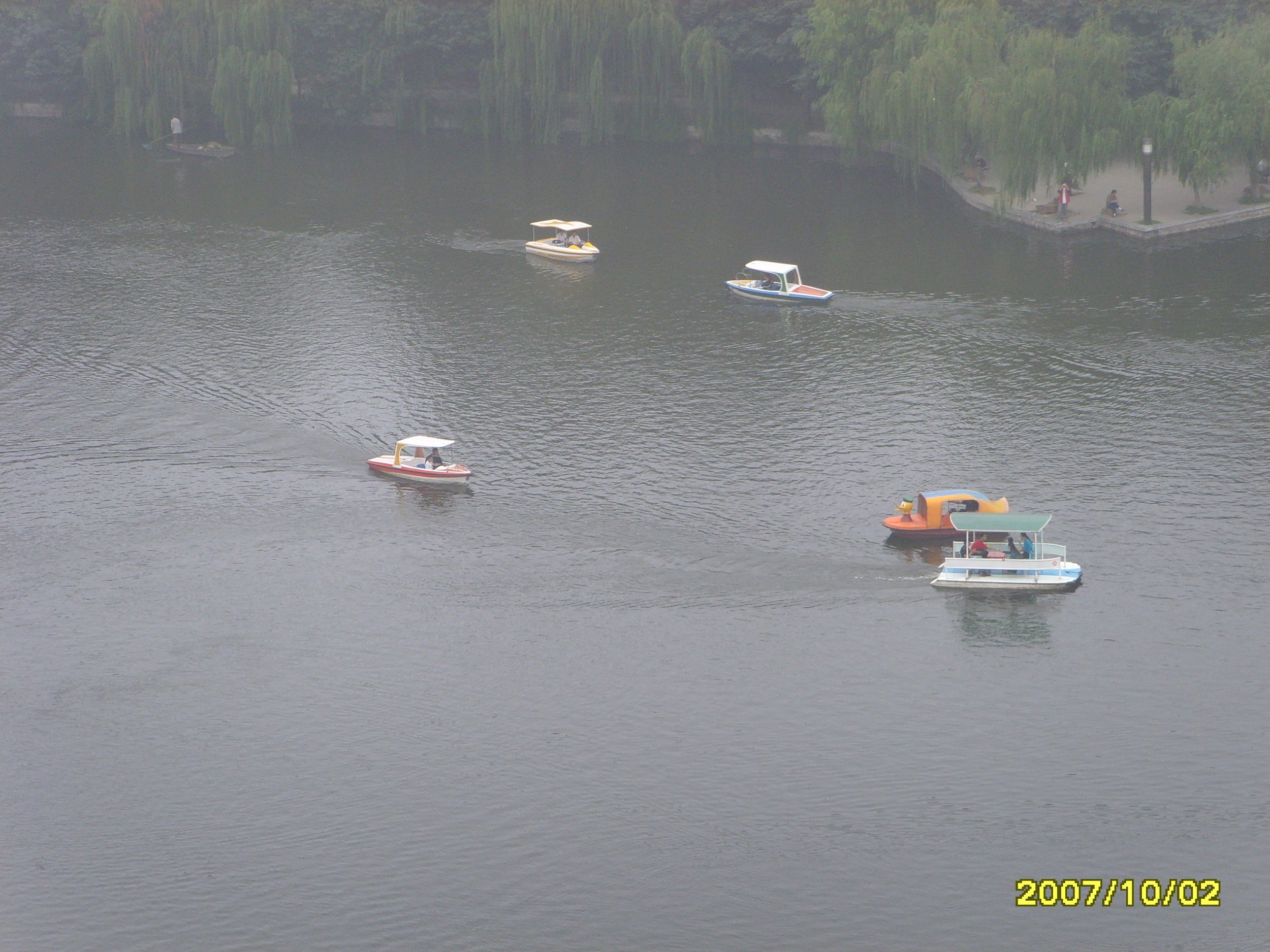 Several small motorboats with covers on a lake, surrounded by trees and a park area with people walking, on a foggy day.