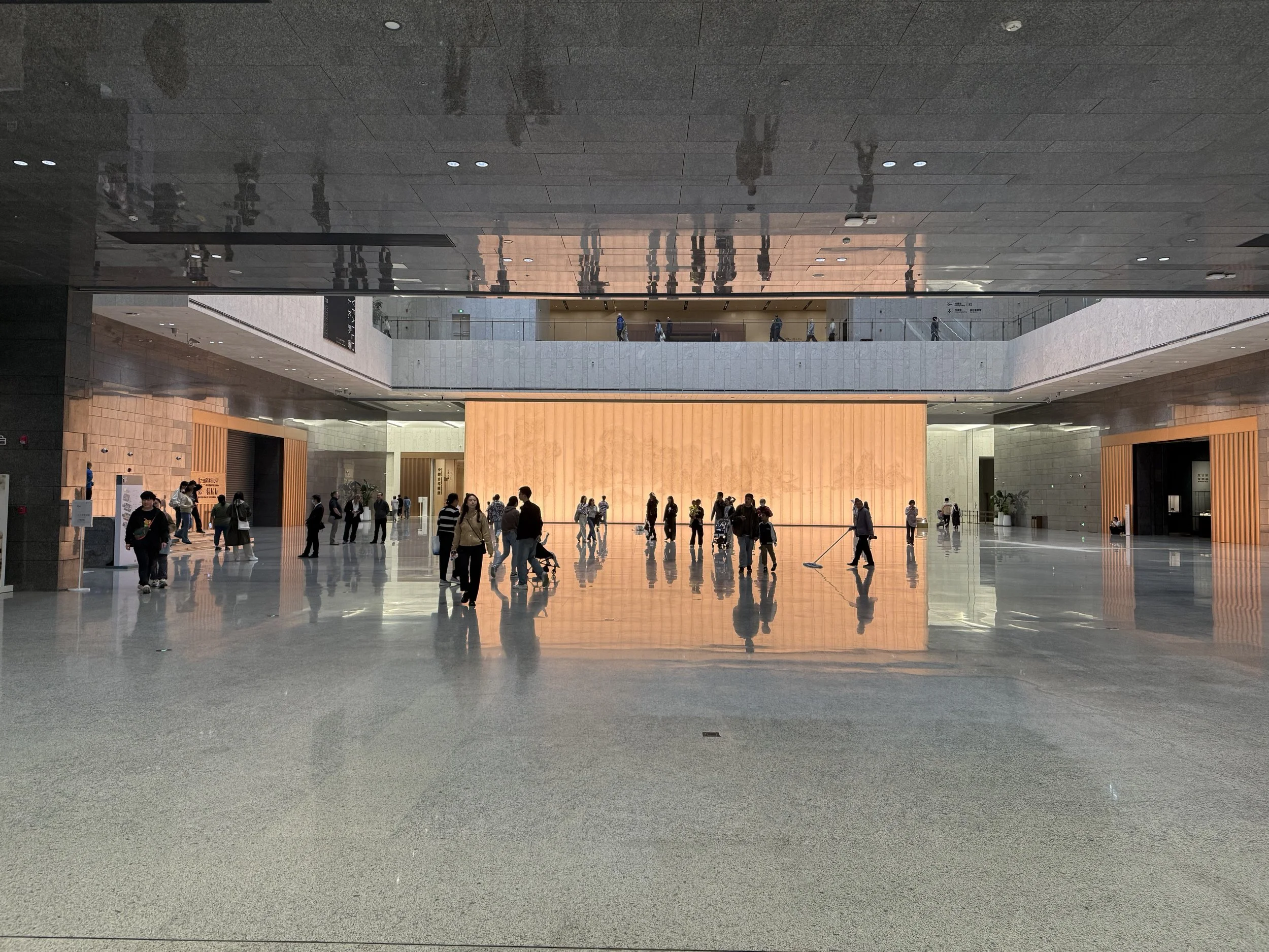 Photo of a spacious modern indoor public space with people walking and standing, high ceiling with lights, and a large warm-colored wall in the background.